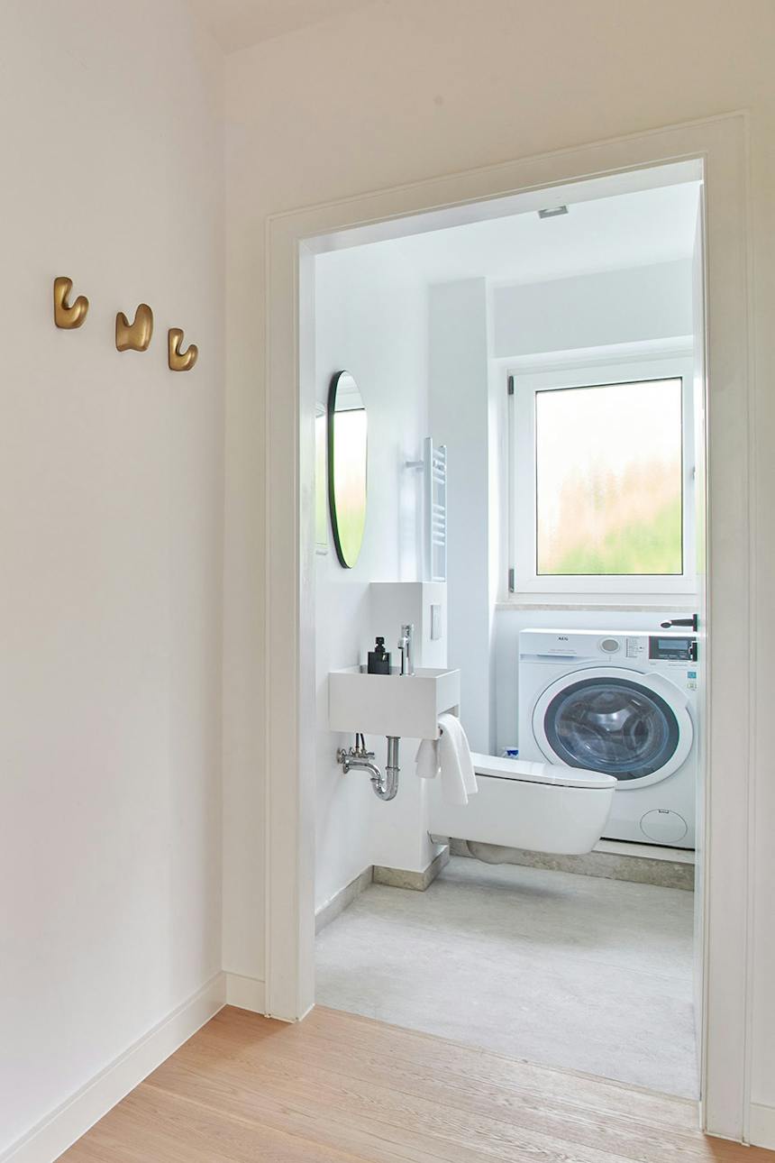 Blick auf ein kleines, helles WC mit Waschmaschine, hängendem WC-Becken und Waschbecken sowie Messing-Wandhaken im Flur.