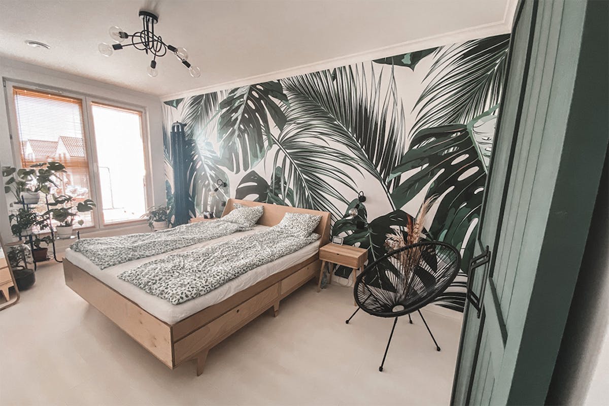 wnętrze sypialni z jasnym drewnianym łóżkiem, dekoracyjną tapetą z motywem roślinnym i czarnym krzesłem ażurowym