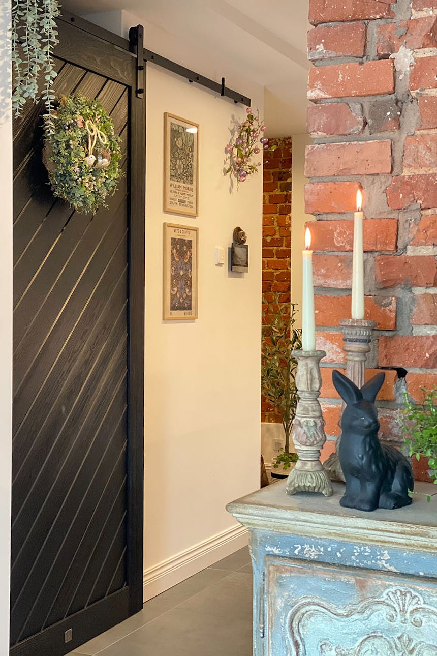 Schiebetür für Speisekammer, schwarze Schiebetür aus Massivholz, dekorativer Kranz an der Tür, roter Backstein an der Wand, dekorative Figuren und Kerzenständer auf der Kommode