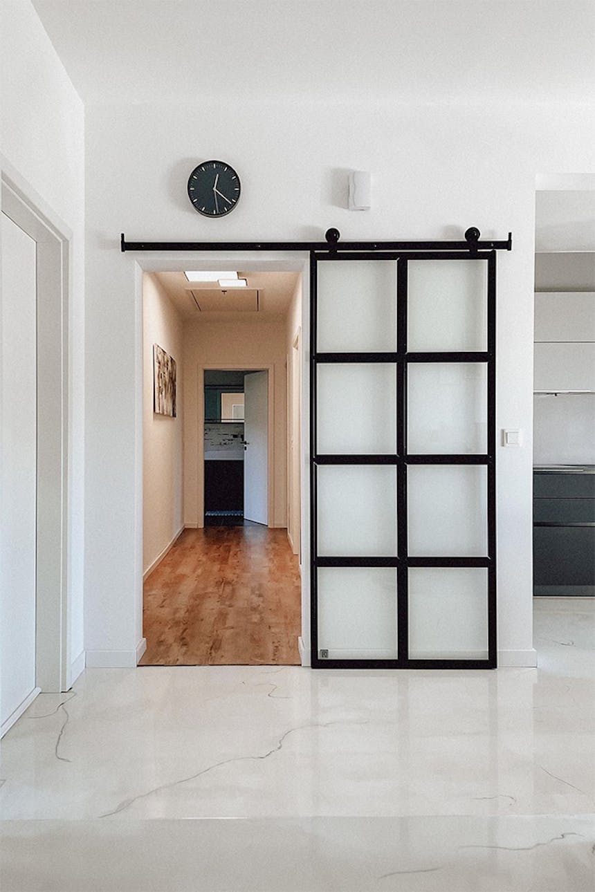 Glasschiebetür, Glastür trennt den Flur, heller Marmorboden, weiße Wände und schwarze Accessoires, schwarze Uhr an weißer Wand, Metalltür mit Glas