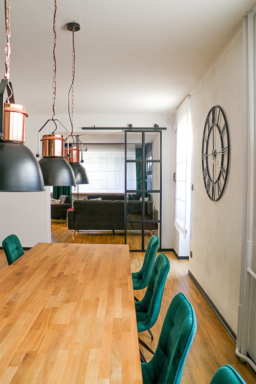 schwarze Metallwanduhr, Uhr im Industriestil, Loftdekor, Metalltür mit Glas, Lofttür, Metallglasschiebetür, schwarze Glastür, grüne Esszimmerstühle, offenes Wohnzimmer zum Esszimmer, industrielle Hängelampen für das Esszimmer, großes hölzernes Esstisch