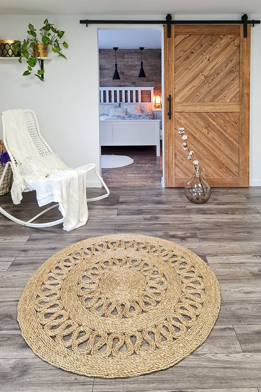 runder, geflochtener Juteteppich, weißer Schaukelstuhl mit Zierkissen, Holzschiebetür im Eingang zum Schlafzimmer, weiße Wände in skandinavischem Interieur, weißes Bett im Schlafzimmer