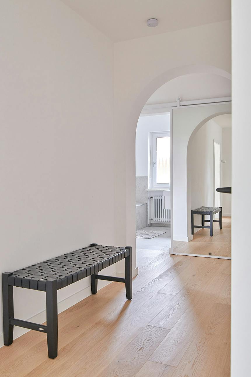 Blick auf einen hellen Flur mit einer schwarzen Bank und einem gewölbten Durchgang, der zu weißen Schiebetüren mit einem Spiegel in voller Größe führt.