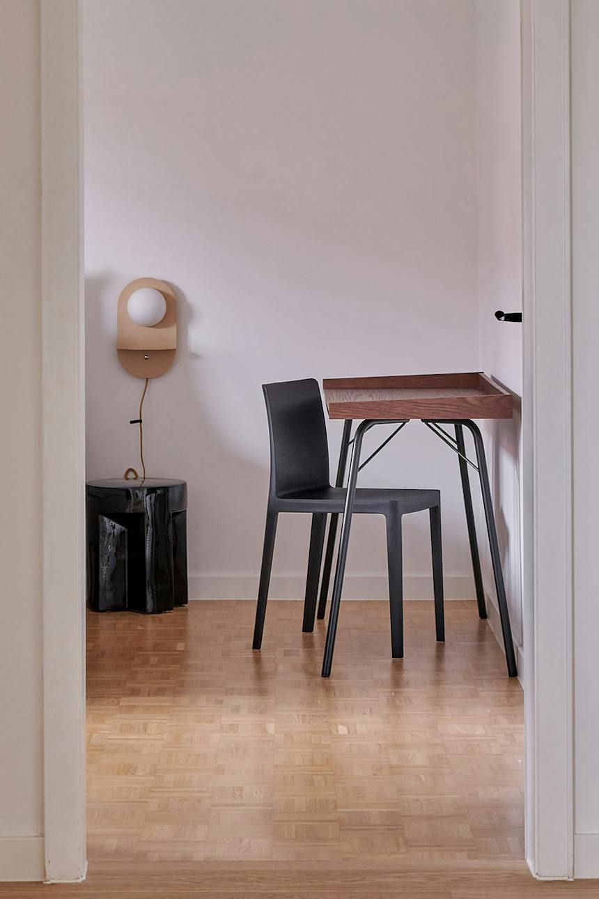 Eine Arbeitsecke mit einem Schreibtisch aus Holz, einem schwarzen Stuhl und einer dekorativen Wandleuchte vor dem Hintergrund einer hellen Wand und eines Parkettbodens.
