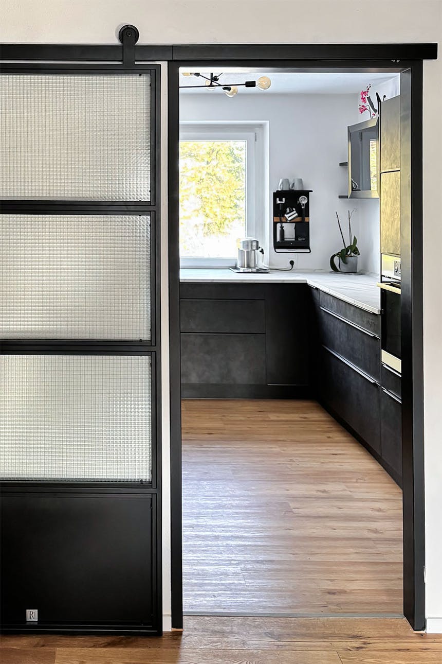 nowoczesna kuchnia z ciemnymi szafkami widoczna przez częściowo otwarte drzwi przesuwne z panelami ze szkła zbrojonego