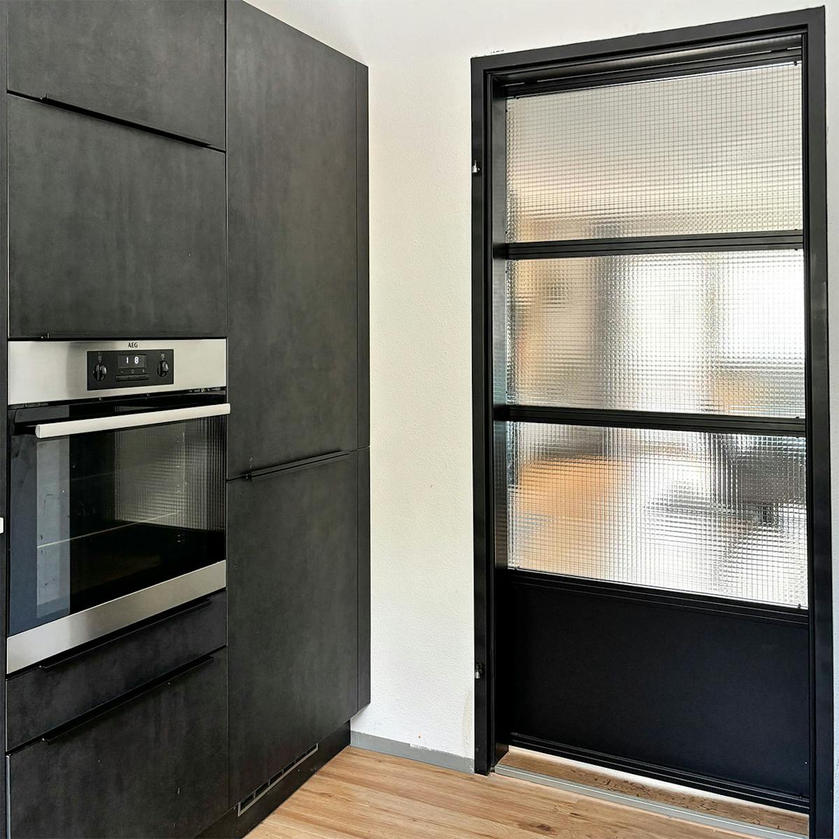 nowoczesna kuchnia z czarnymi szafkami i wbudowanym piekarnikiem obok przesuwnych drzwi ze stali i szkła zbrojonego