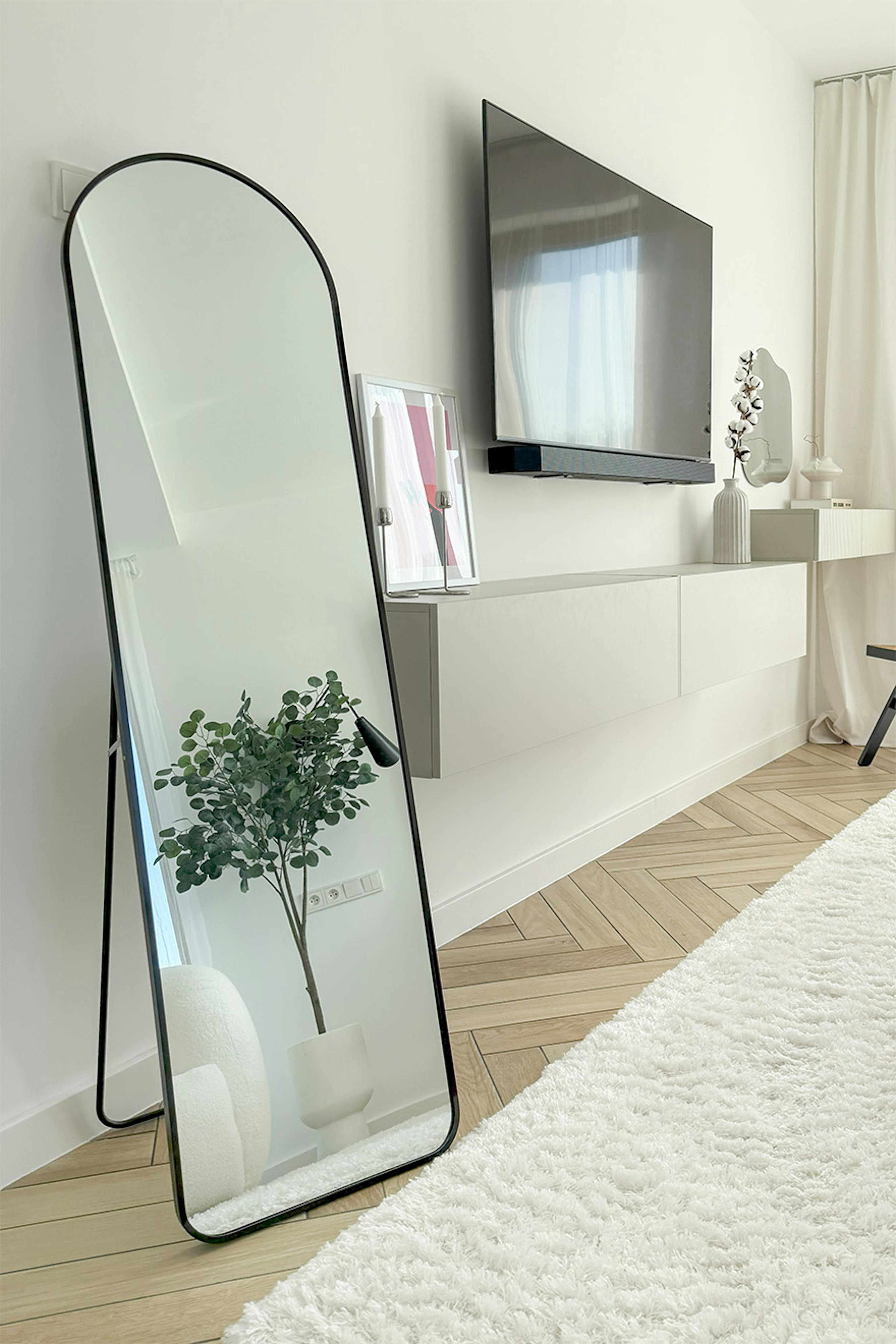 un coin dans une chambre moderne avec un grand miroir noir arqué appuyé contre un mur blanc, une télévision murale et une console suspendue