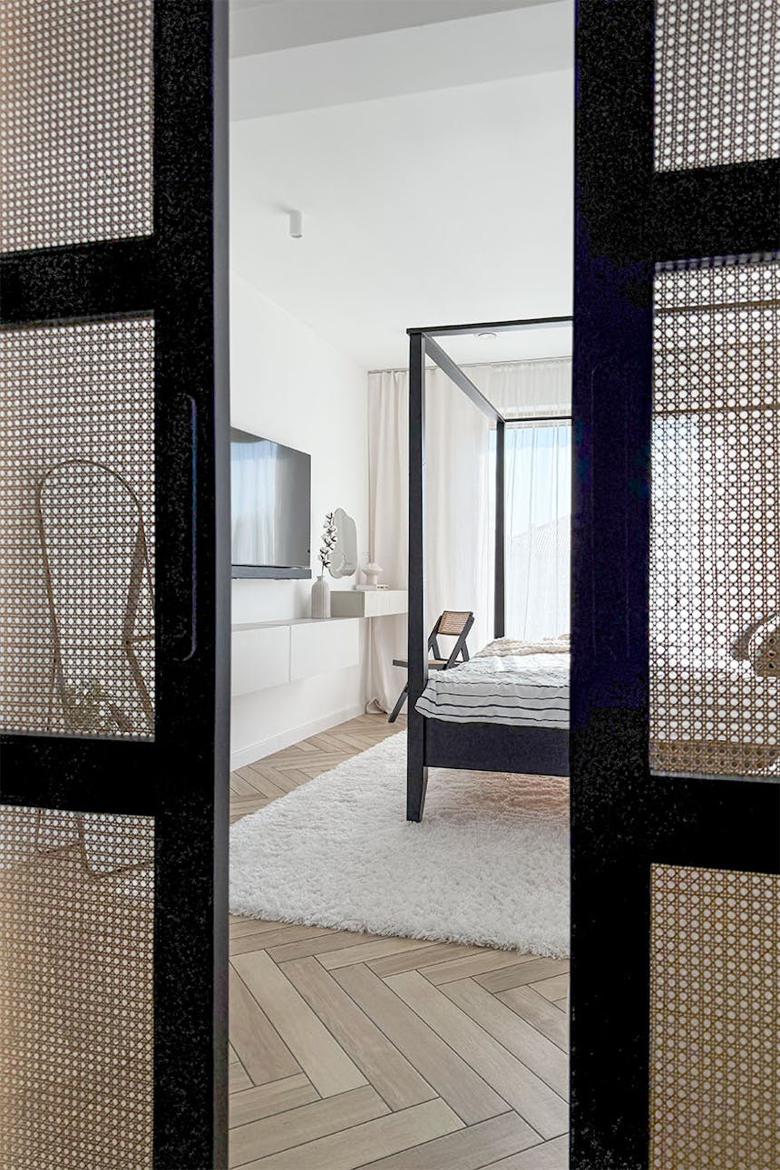 Blick durch schwarze Rattan-Schiebetüren in minimalistisches Schlafzimmer mit schwarzem Himmelbett, weißen dünnen Vorhängen und hellem Fischgrätholzboden