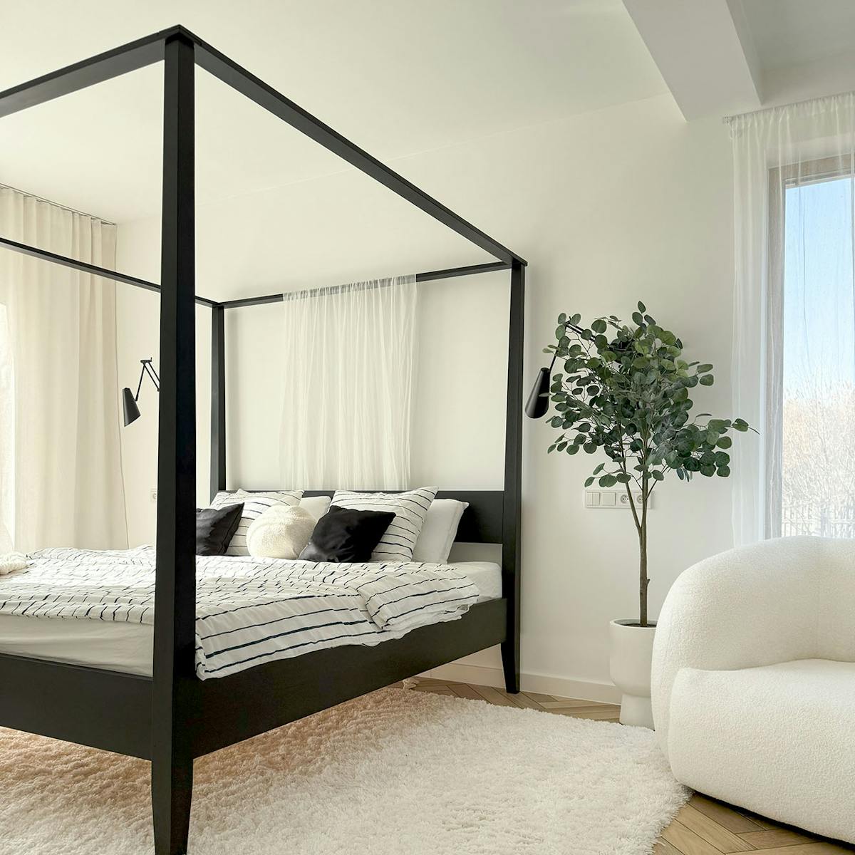 helles Schlafzimmer mit großem schwarzen Himmelbett, gestreifter Bettwäsche, großem weißen Teppich und hoher Pflanze in weißem Topf