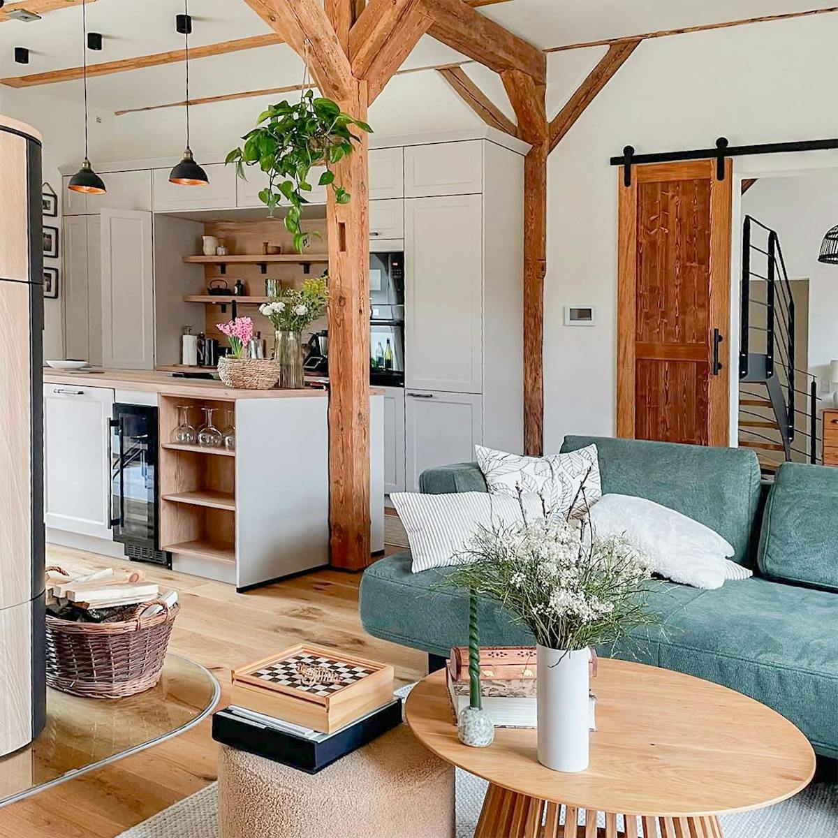 Offener Wohn- und Küchenbereich in einem renovierten Stadl, mit sichtbaren Holzbalken, einem grünen Sofa, modernen weißen Küchenmöbeln und rustikalen Details.