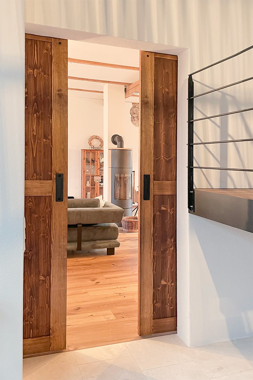 Open wooden sliding barn doors leading to a bright living room with wooden flooring, freestanding stove and dark sofa.