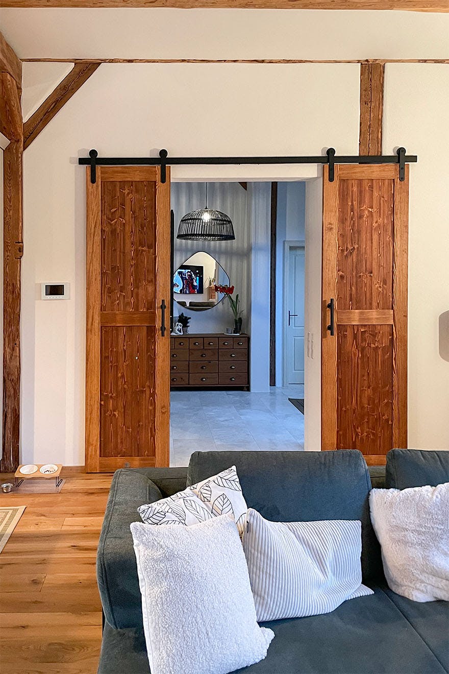 View of the open, wooden sliding barn door, behind which you can see the hall with a mirror and a chest of drawers. In the foreground, a fragment of a dark sofa with cushions.