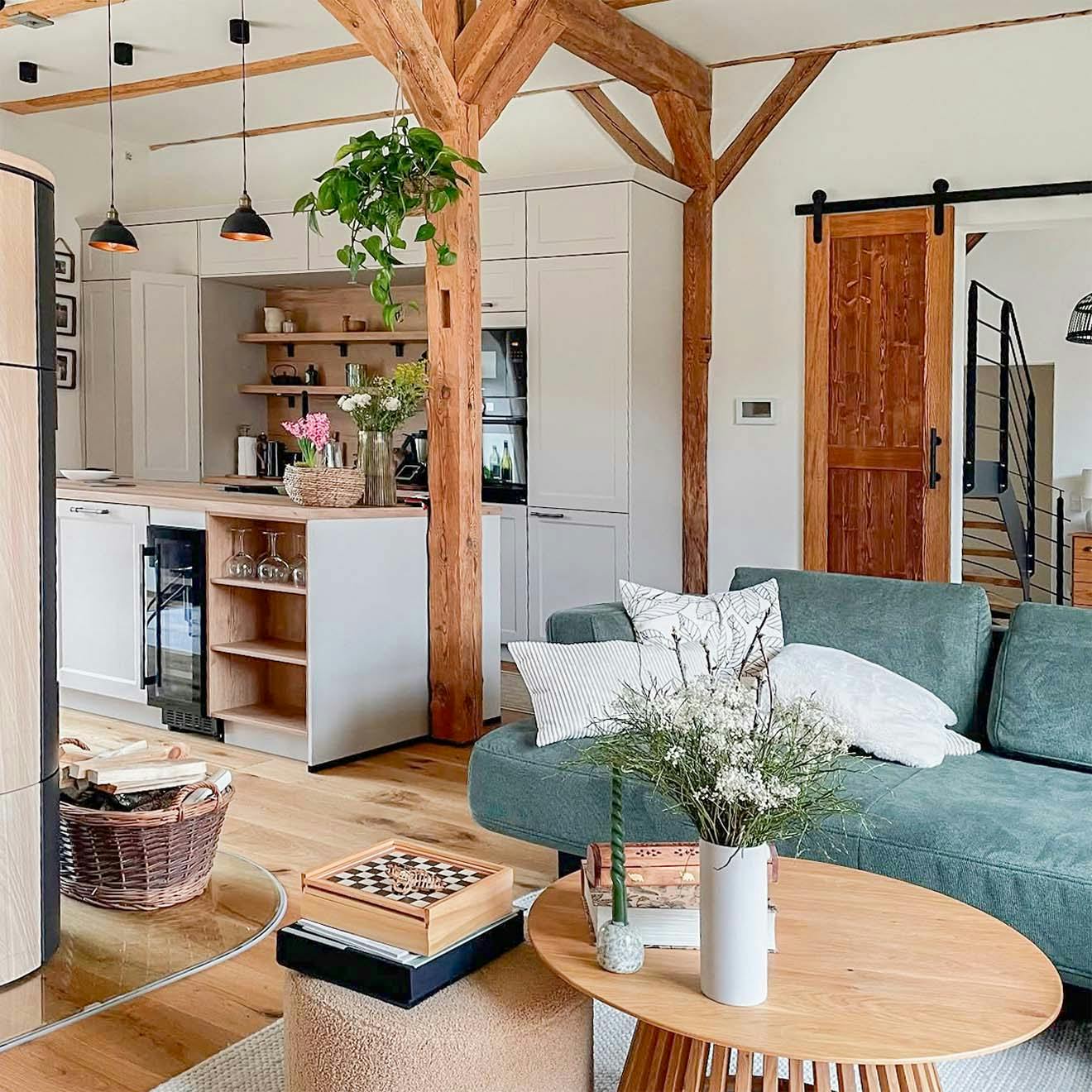 Open-space living room and kitchen in a restored barn, with exposed wooden beams, a green sofa, modern white kitchen units and rustic details.