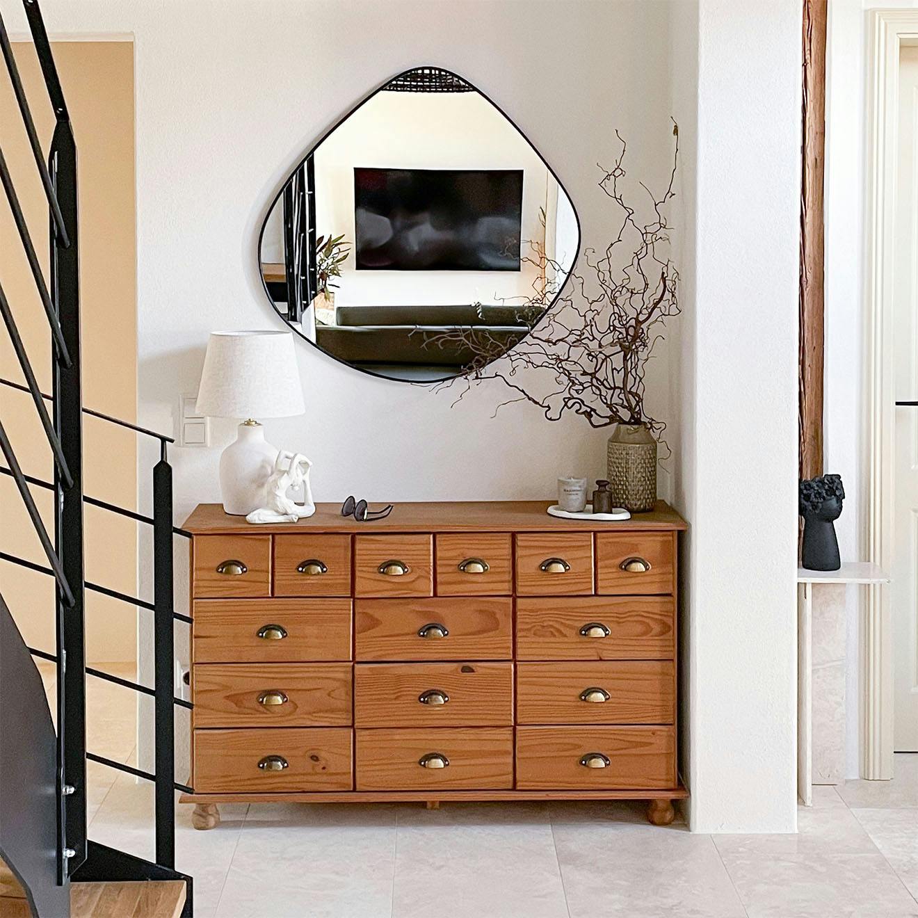 Rustic chest of drawers with numerous drawers and brass handles, under a modern, irregularly shaped mirror, in a bright hall.