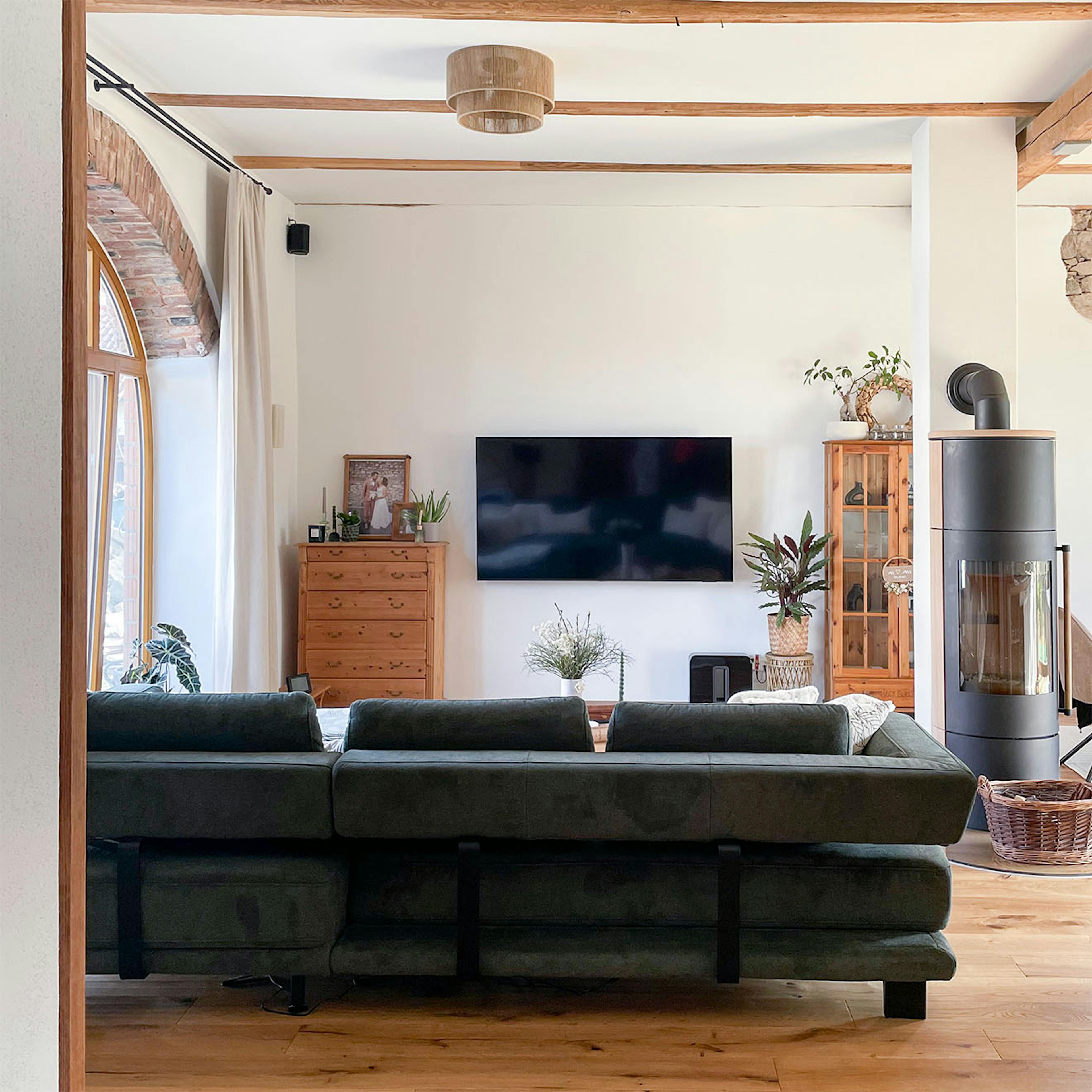 Séjour spacieux et lumineux dans une grange restaurée, avec télévision murale, grand canapé sombre, plafond à poutres apparentes et poêle indépendant.