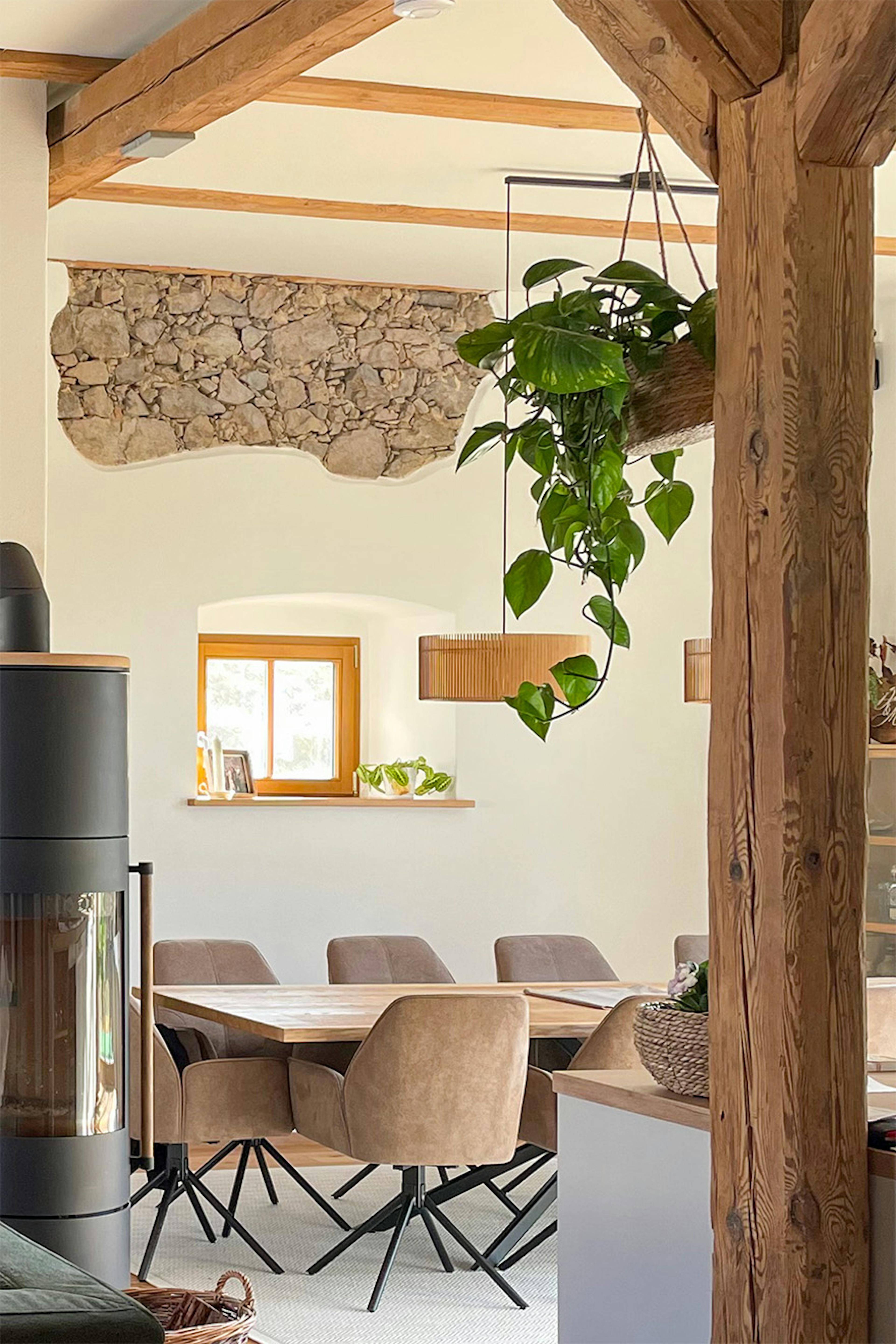 Salle à manger dans une maison de campagne moderne avec table en bois, chaises beiges, plantes suspendues et fragment de mur en pierre.