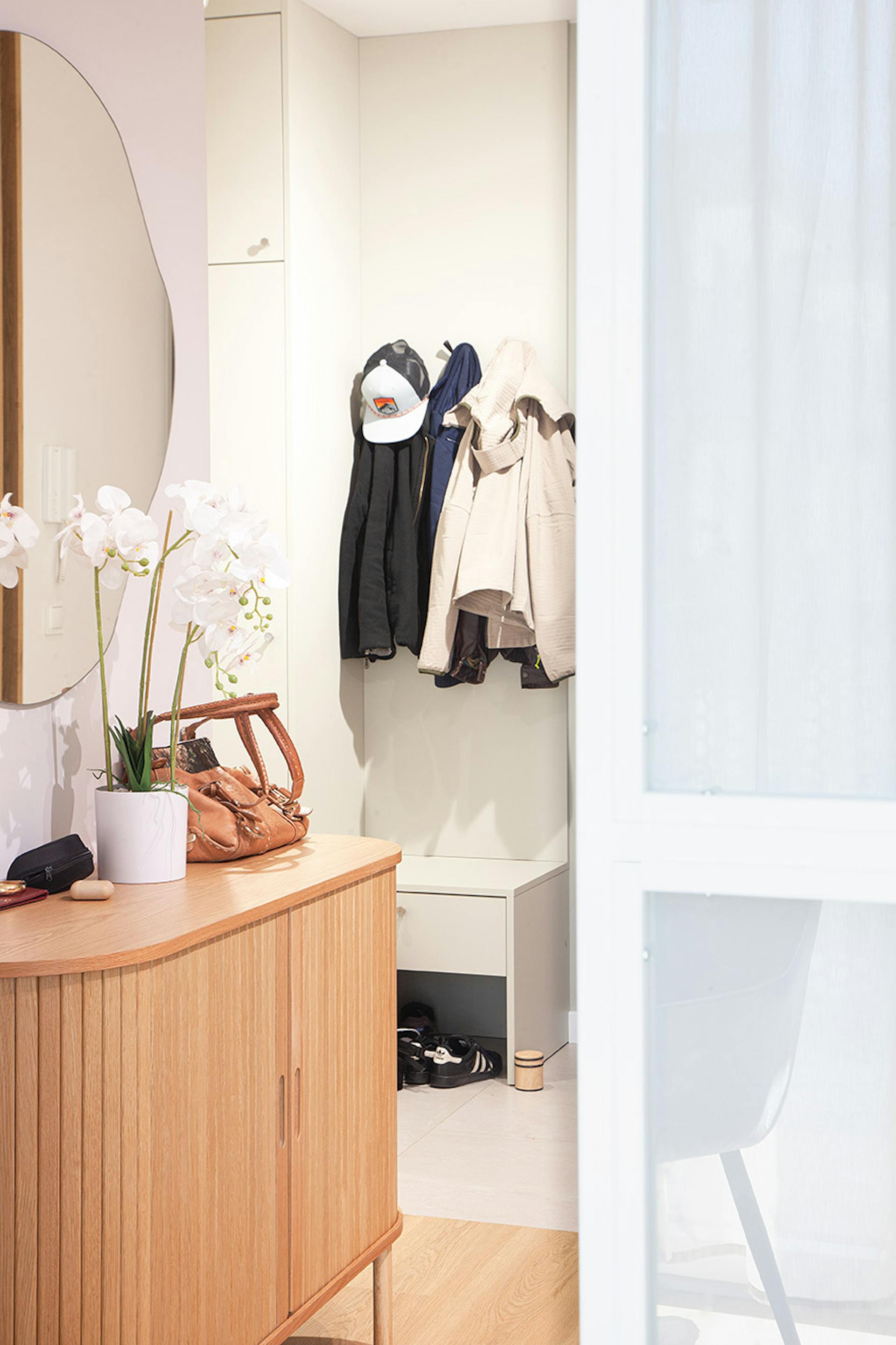 Korytarz wejściowy z drewnianą komodą, okrągłym lustrem i białymi orchideami, w tle wieszak z kurtkami i szafka na buty, częściowo widoczny przez białe drzwi przesuwne.