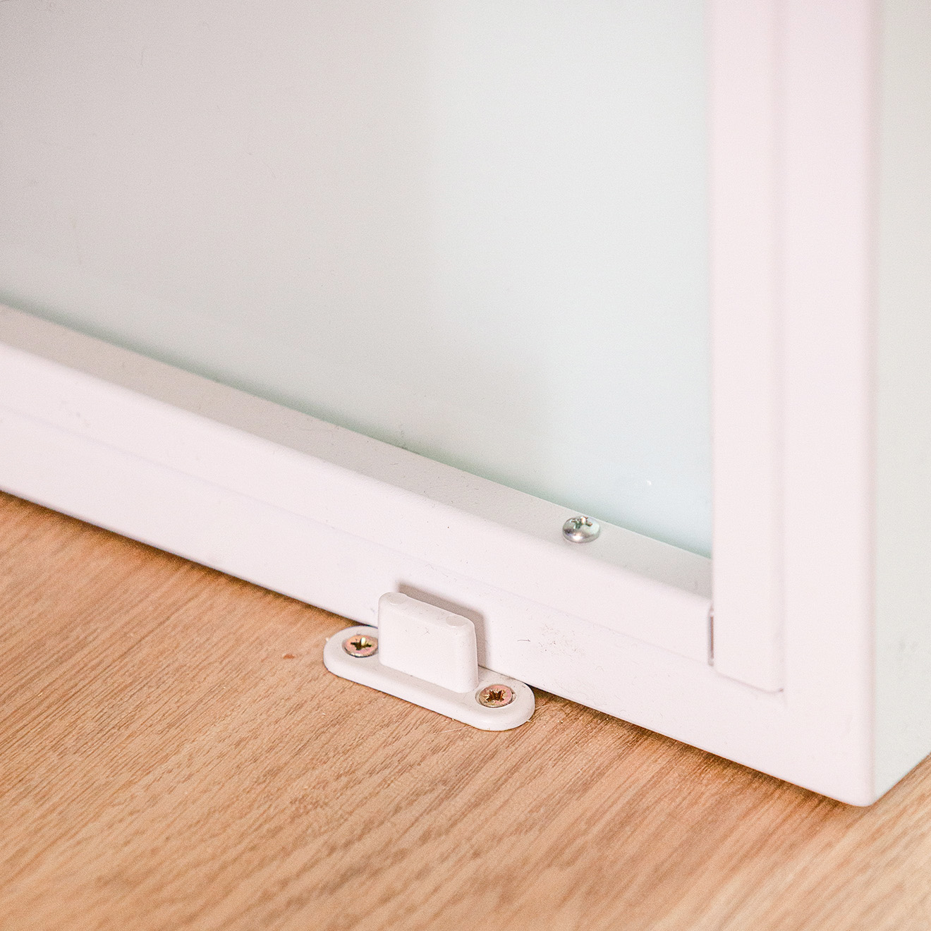 Detal białego prowadnika podłogowego przy dolnej ramie białych drzwi przesuwnych, przymocowanego do drewnianej podłogi.