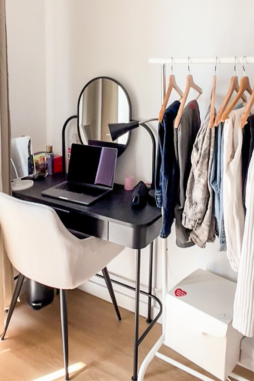 Czarne biurko z okrągłym lustrem, obok tapicerowane krzesło i stojący wieszak na ubrania we wnętrzu z jasnymi ścianami i drewnianą podłogą.