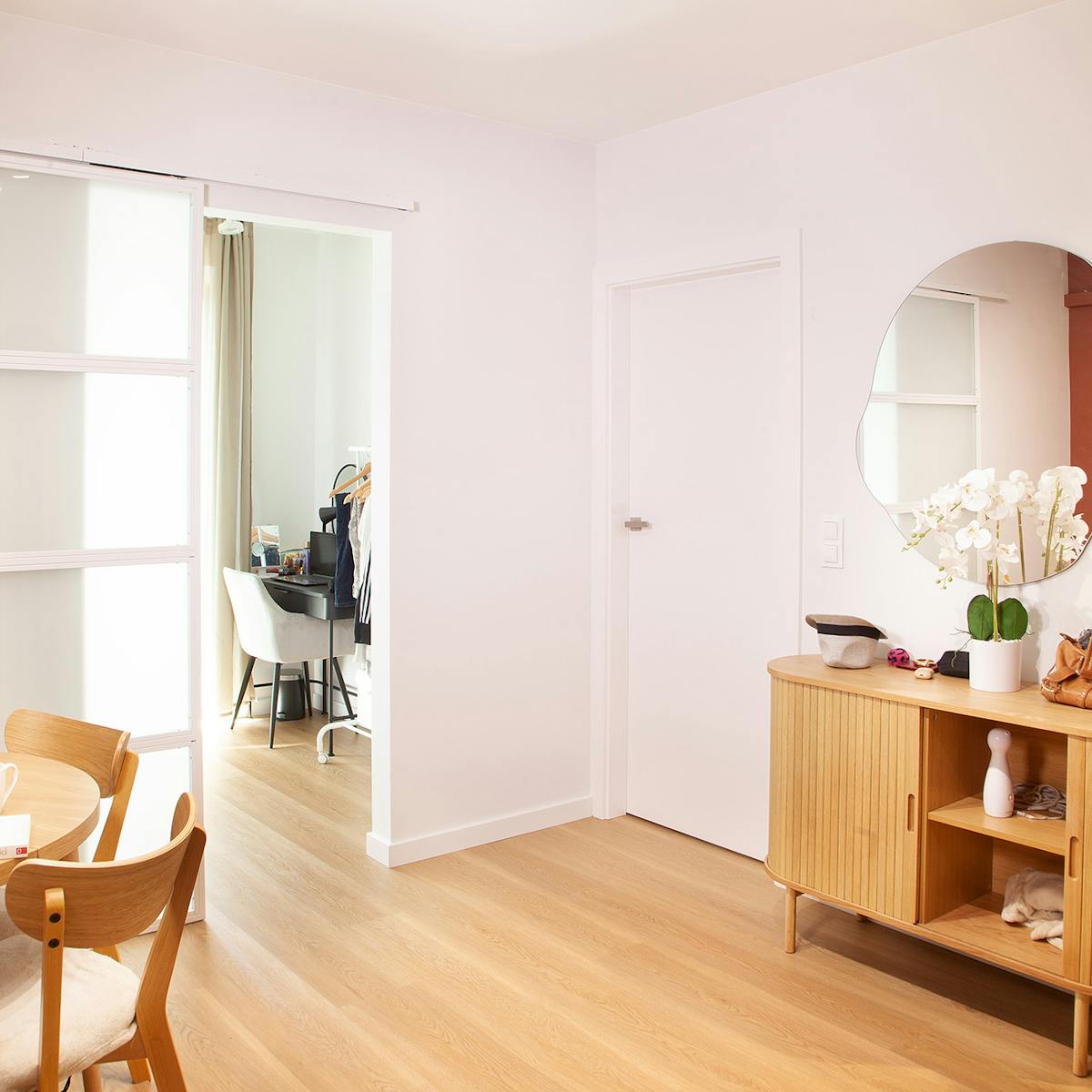Blick auf den offenen Ess- und Eingangsbereich mit einer Kommode aus Holz und einem runden Spiegel an der Wand sowie weißen Schiebetüren, die zum Schlafzimmer führen.
