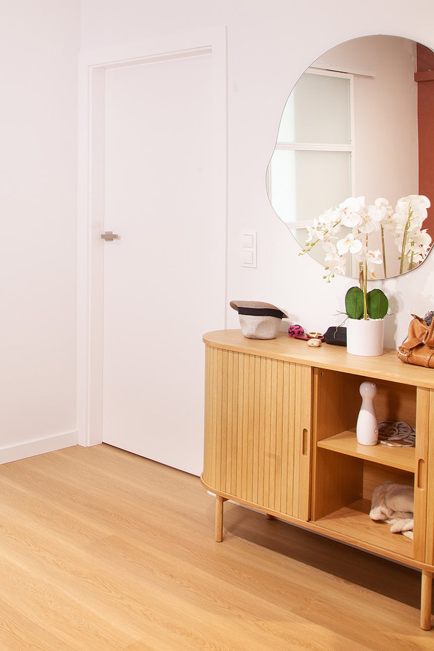 Heller Flur mit einer Holzkommode mit abgerundeten Ecken und offenen Regalen, auf der weiße Orchideen stehen; ein Spiegel mit unregelmäßiger Form an der Wand über der Kommode.