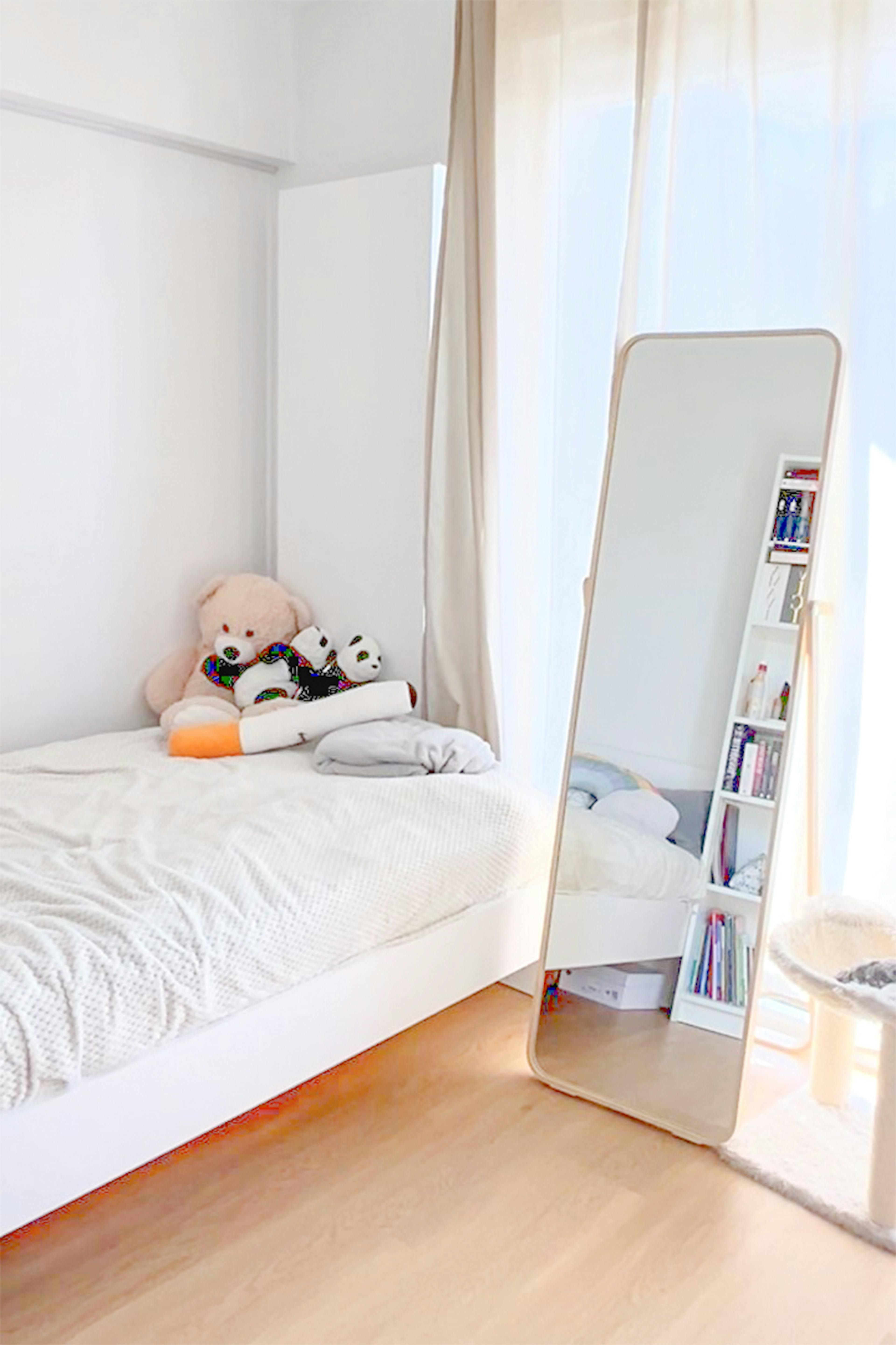 Chambre lumineuse avec un lit blanc sur lequel sont posés des ours en peluche, et un grand miroir sur pied reposant sur un parquet en bois près de la fenêtre.