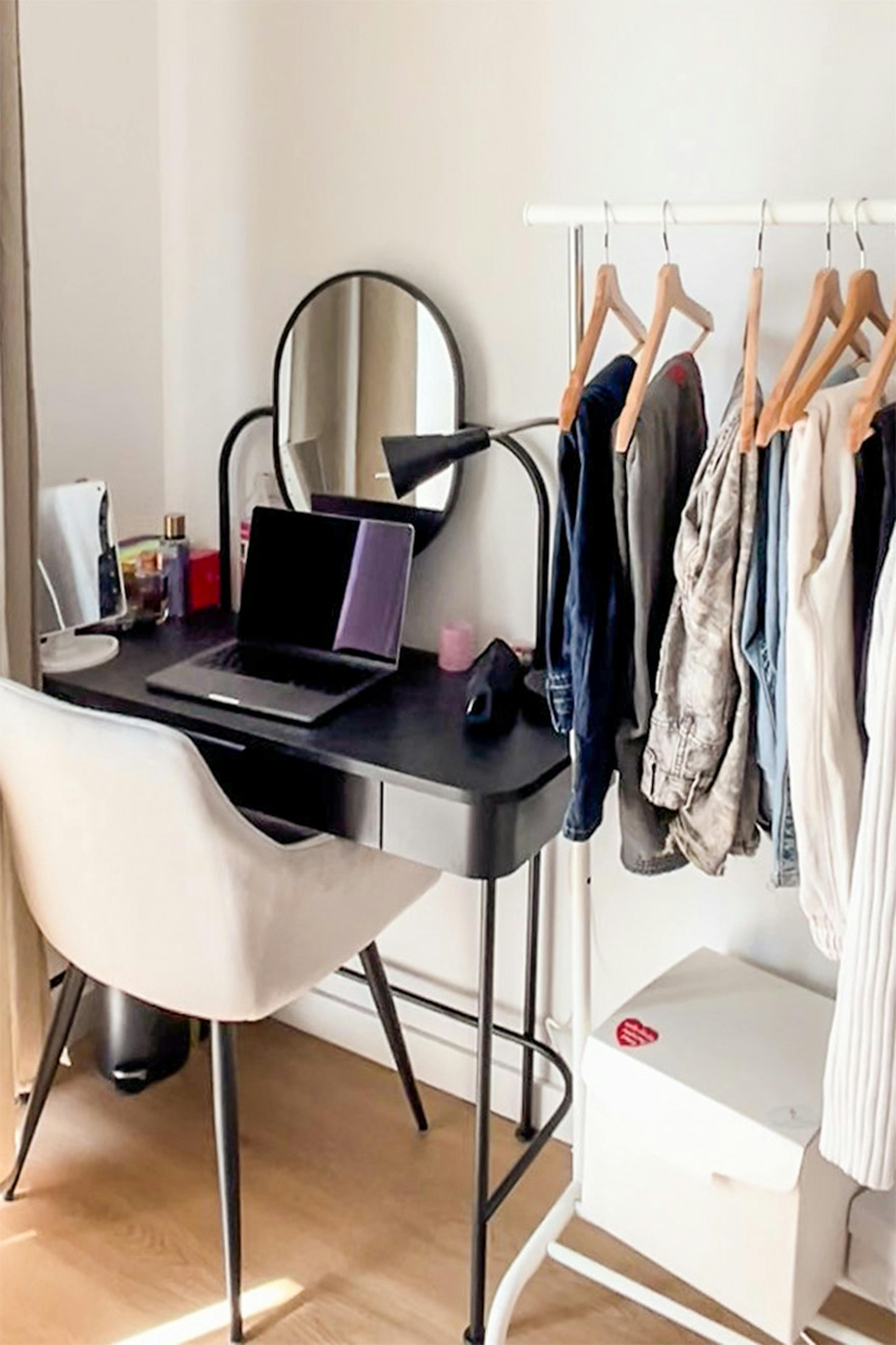Bureau noir avec un miroir rond, à côté d'une chaise tapissée et d'un portemanteau sur pied dans un intérieur aux murs clairs et au parquet en bois.