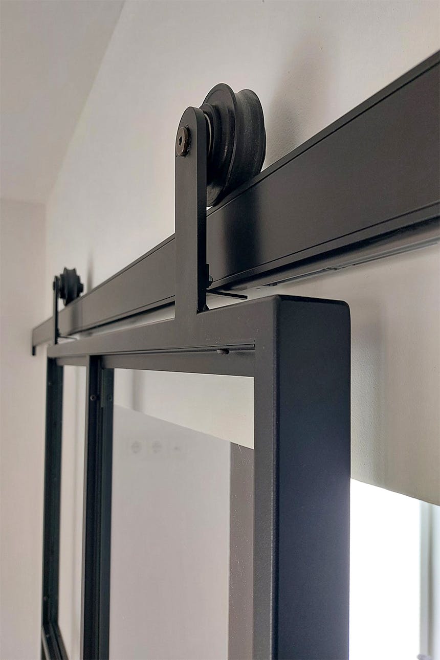 Nahaufnahme des Schiebemechanismus einer schwarzen Industrietür aus Metall und Glas, wobei Führungsschiene und Rolle sichtbar sind.