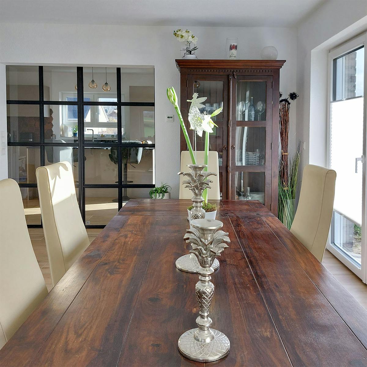 Ein Esstisch aus Holz mit Dekorationen, im Hintergrund eine klassische Vitrine und eine schwarze Industrietür, die zur Küche führt.