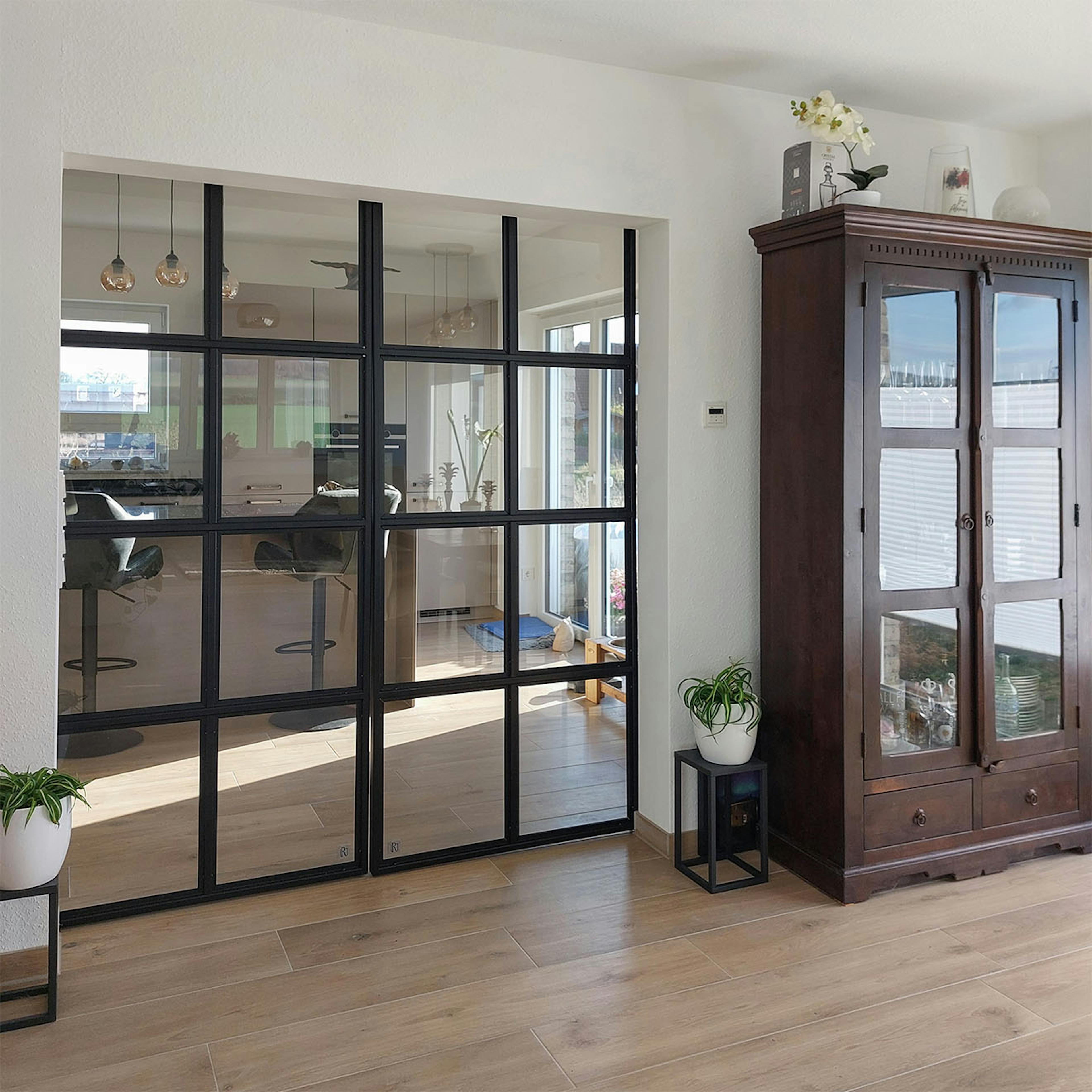 Des portes en verre industrielles séparent le salon de la cuisine par un îlot, à côté duquel se trouve une vitrine sombre et sculptée de style classique.