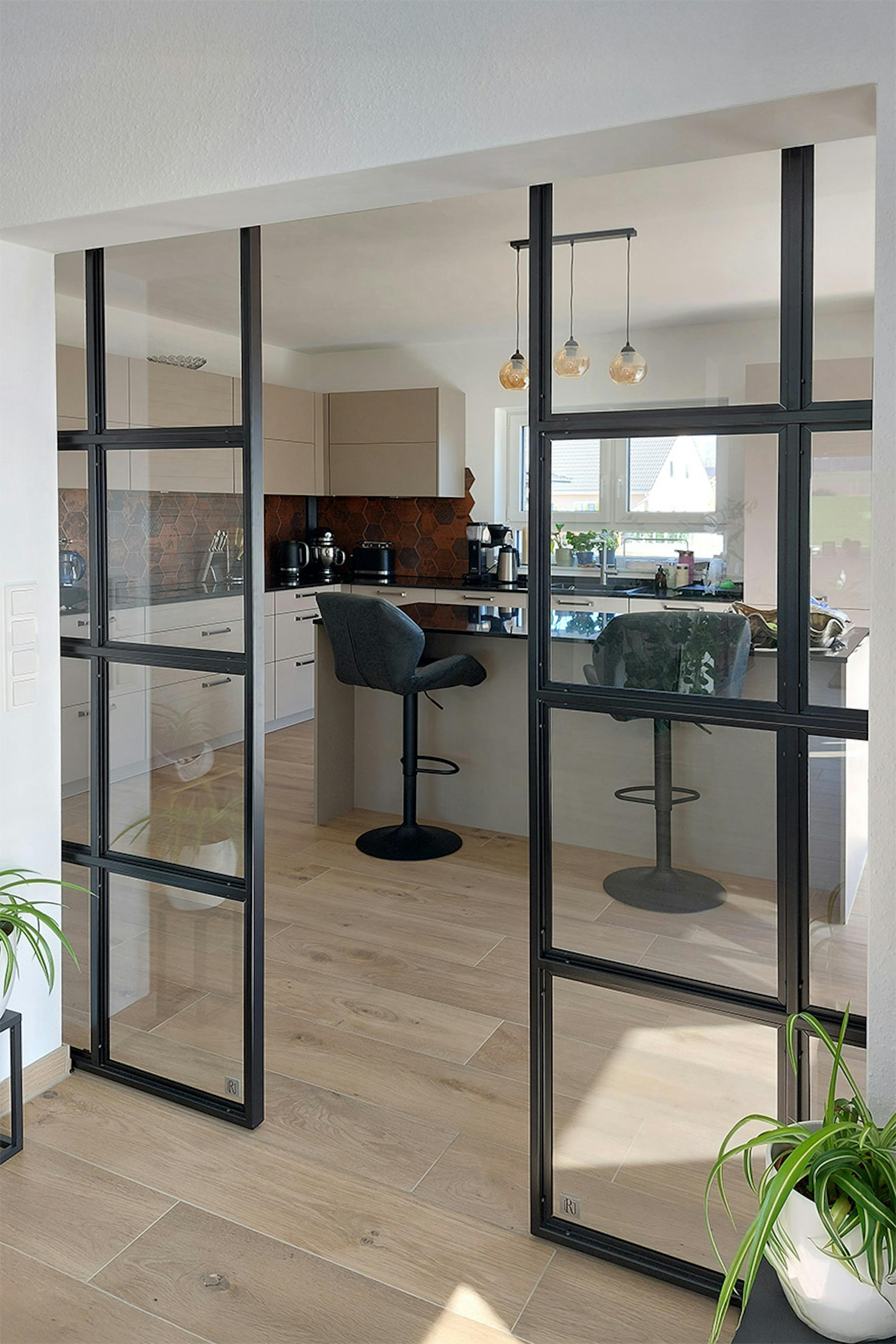 Le salon s'ouvre sur la spacieuse cuisine par des portes industrielles noires. On aperçoit la cuisine, avec son îlot central, ses façades beiges et son carrelage cuivré.