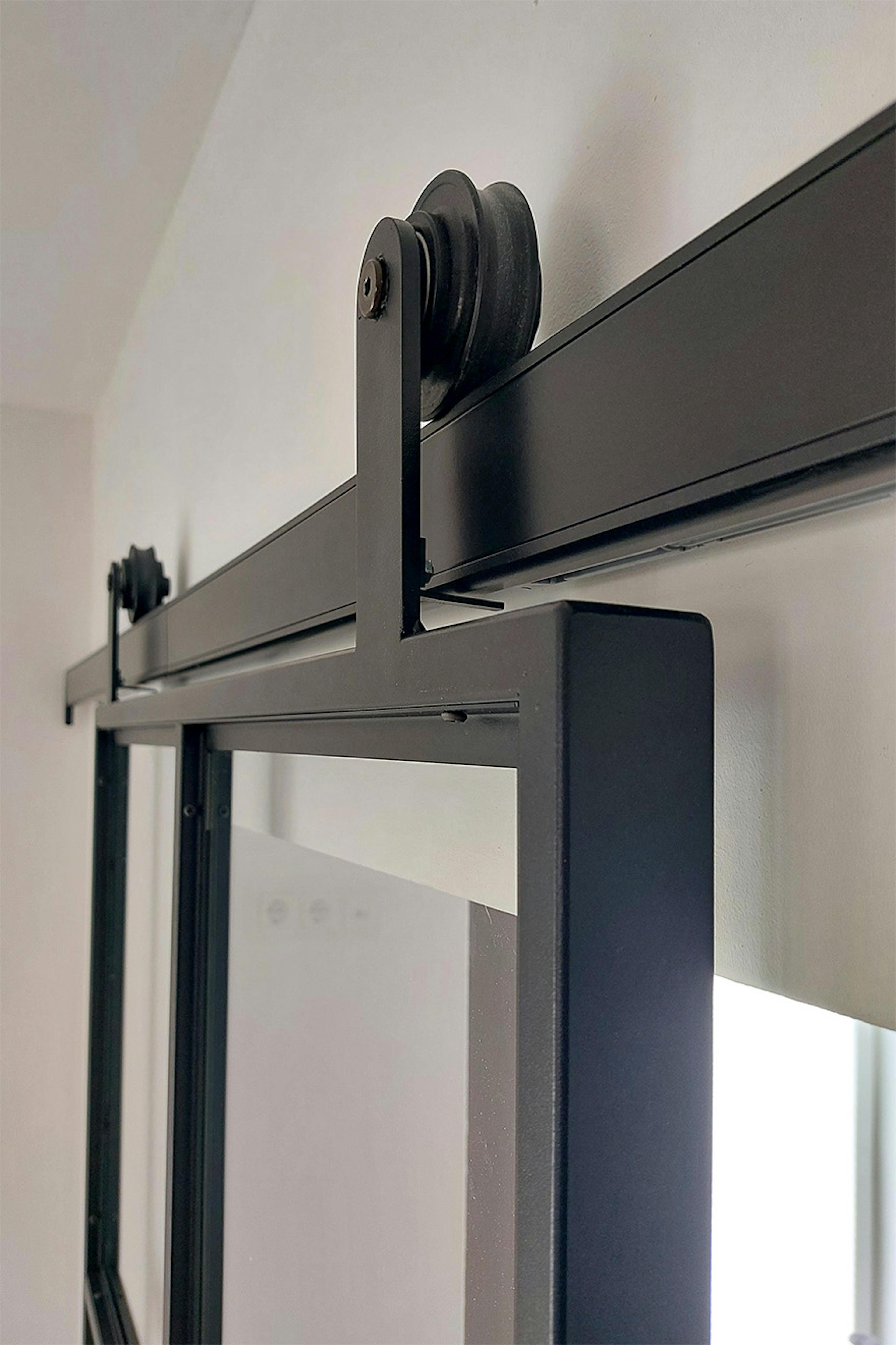 Gros plan du mécanisme coulissant d'une porte industrielle noire en métal et verre, avec le rail et le rouleau visibles.