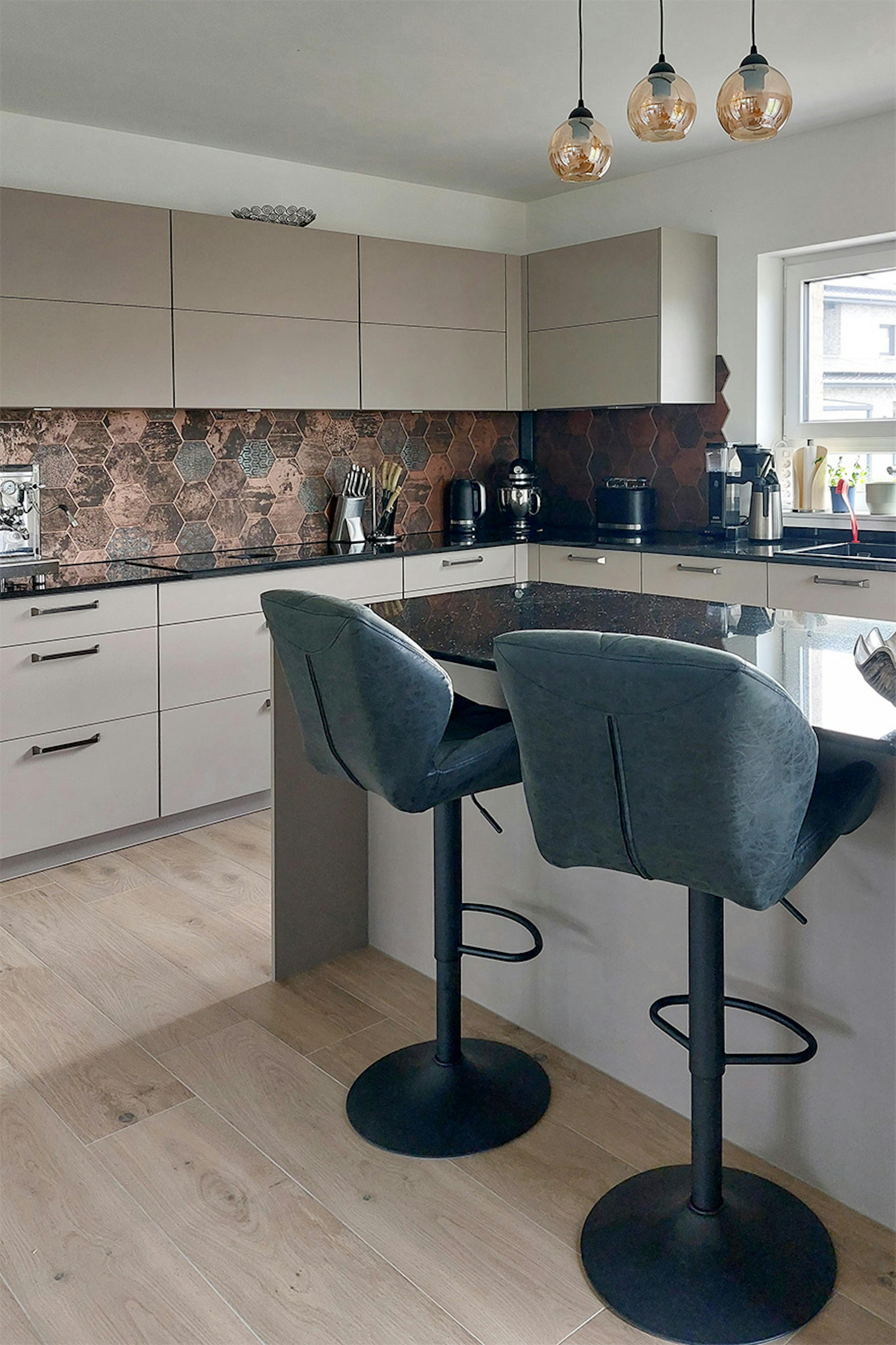 Cuisine avec îlot, façades beiges, carrelage en cuivre, tabourets de bar noirs et lampes suspendues - design moderne et fonctionnel.