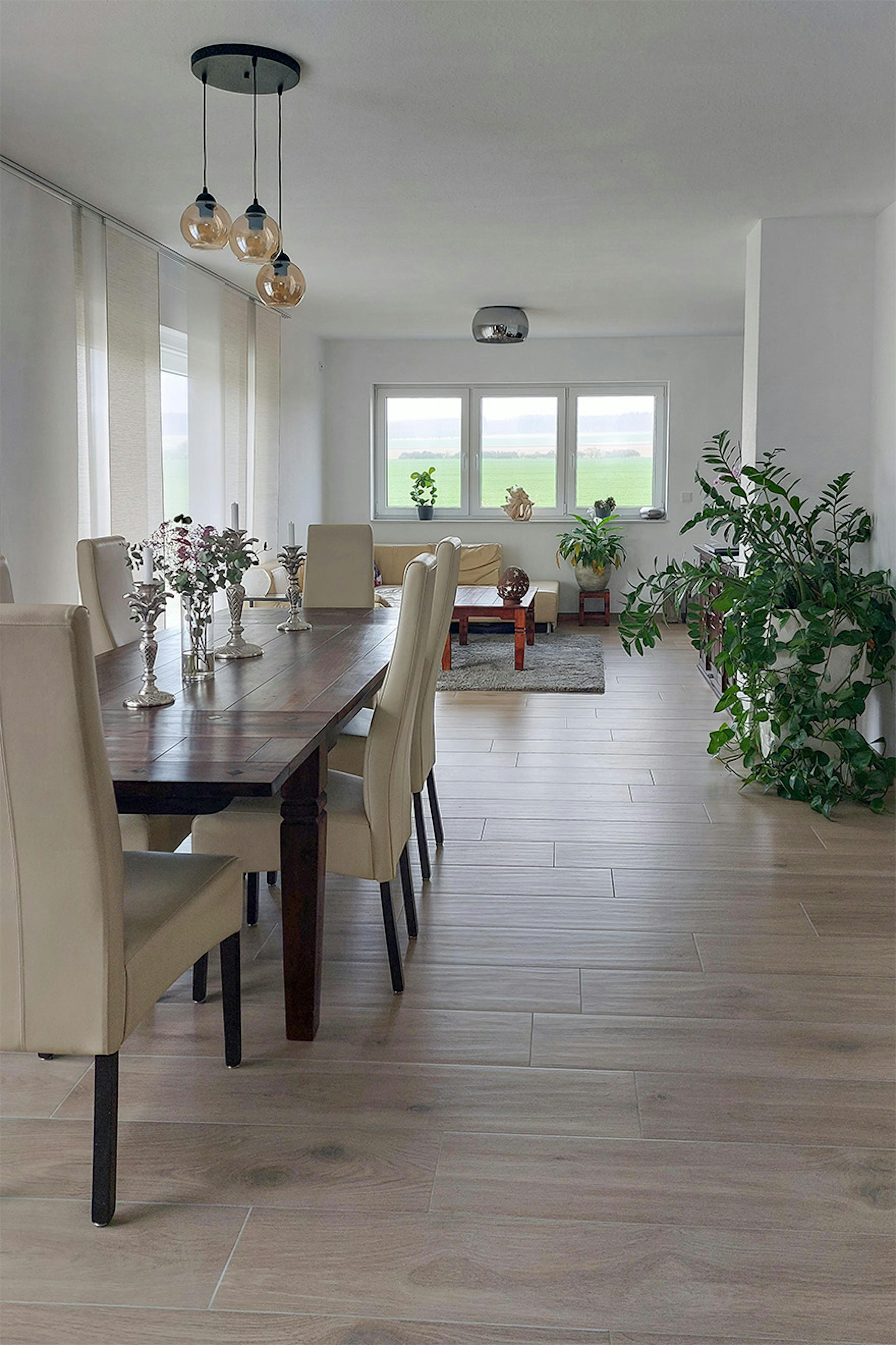 Une salle à manger lumineuse avec une longue table en bois et des chaises crème, au fond un grand salon avec un canapé beige et des fenêtres donnant sur la verdure.