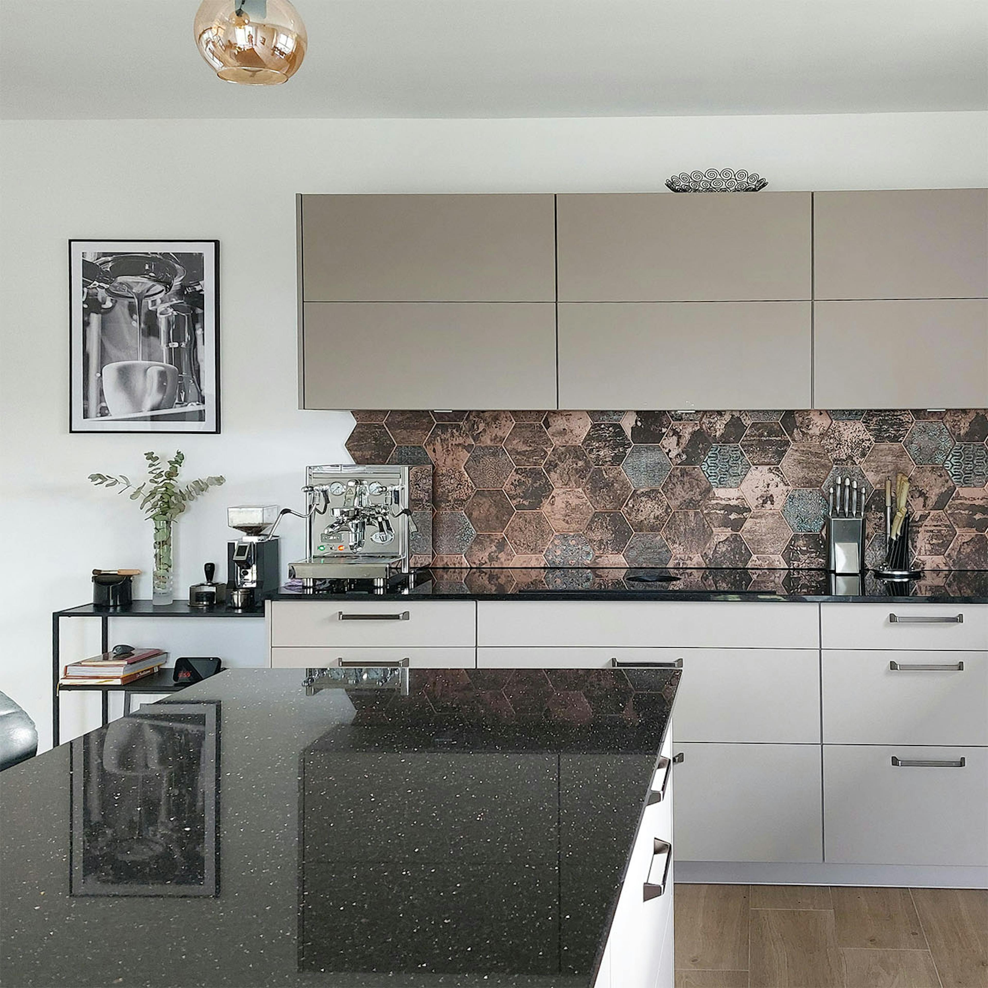 Cuisine moderne avec armoires beiges, comptoir foncé, carreaux hexagonaux en cuivre et cafetière argentée.