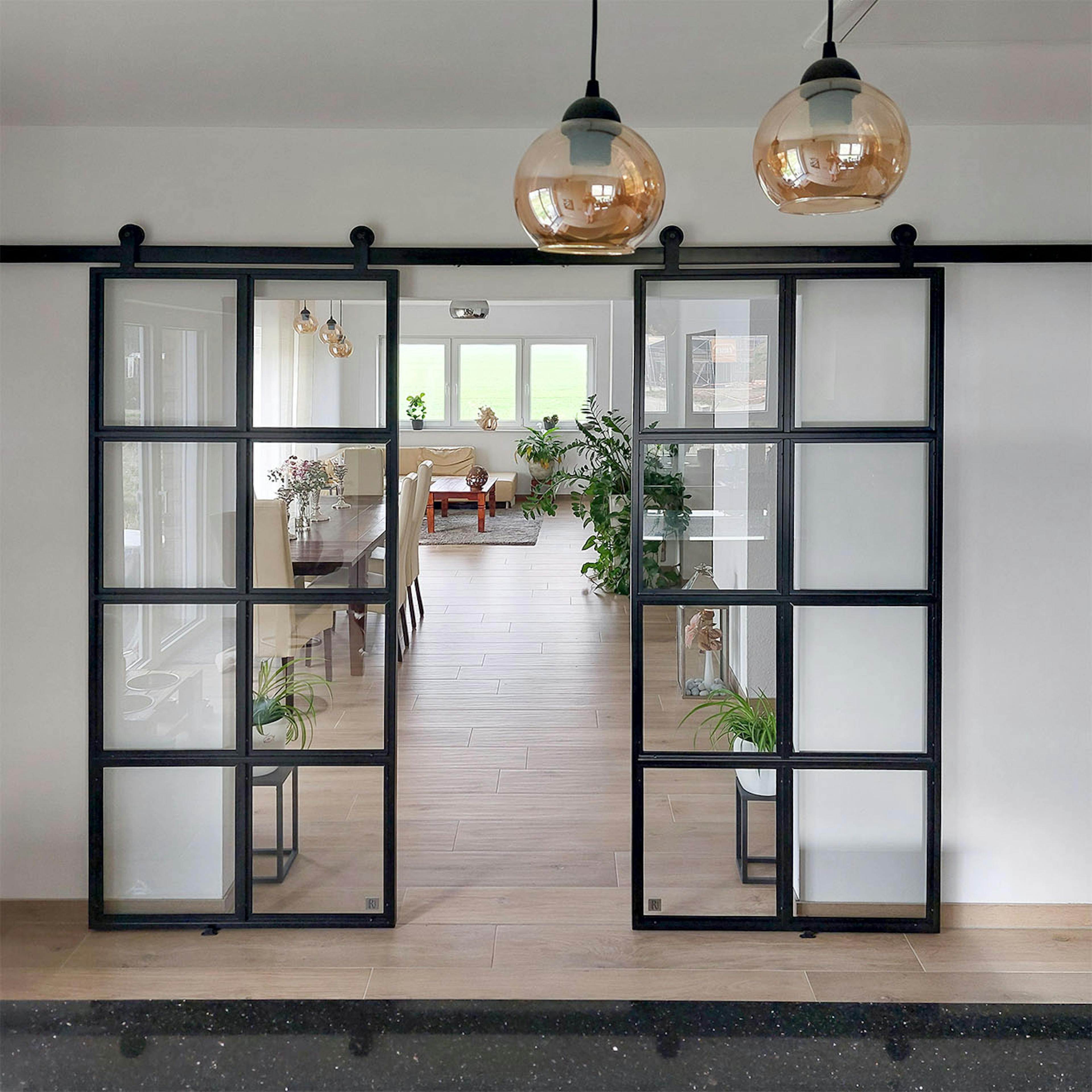 Des portes coulissantes industrielles noires séparent la cuisine du salon ; un intérieur moderne avec des éléments classiques et de la lumière naturelle.