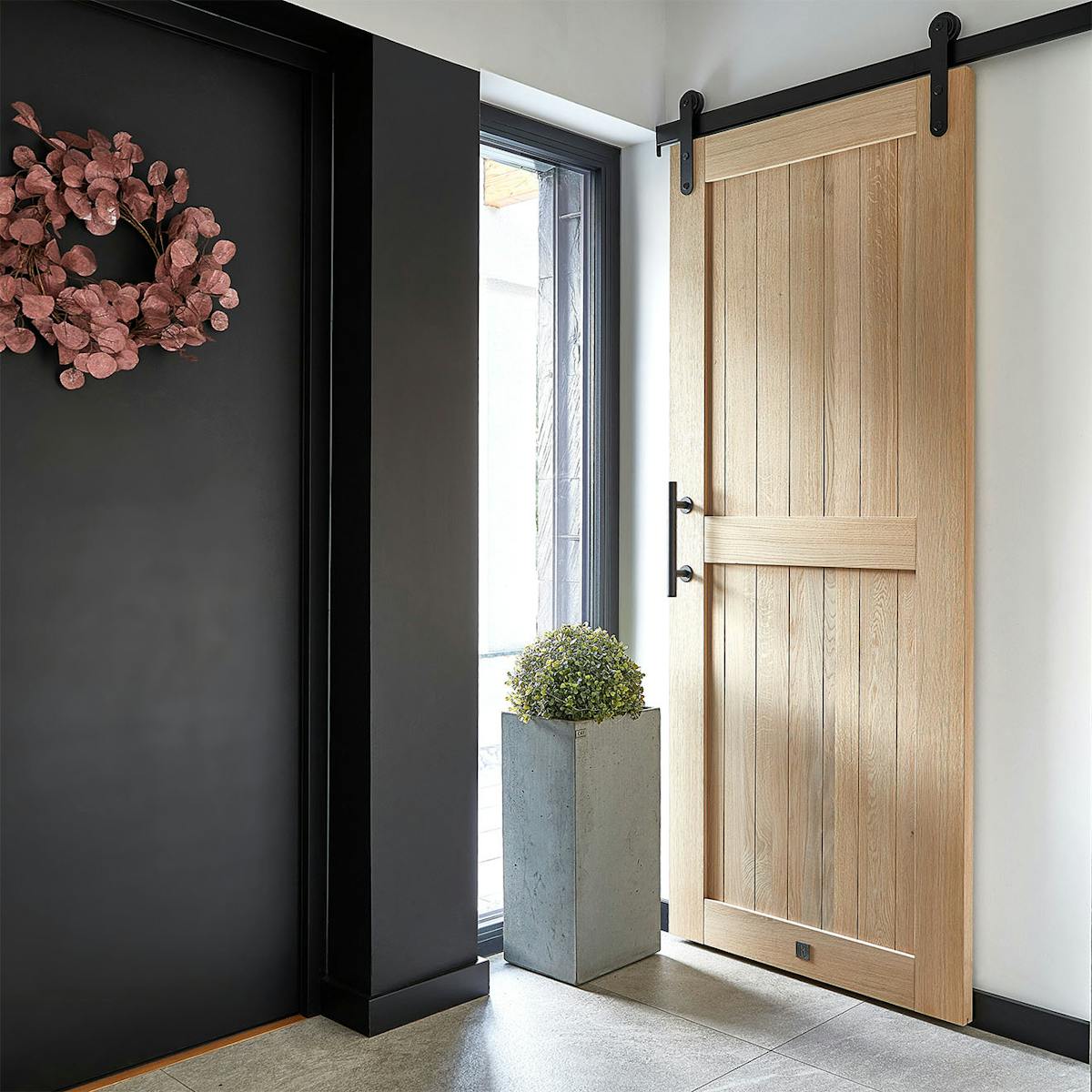 Nowoczesny wiatrołap z dębowymi drzwiami przesuwnymi w stylu barn doors. Jasne, drewniane drzwi z czarnym systemem przesuwnym, obok nich betonowa doniczka i wieniec na ciemnej ścianie, tworzą elegancką aranżację przedpokoju.
