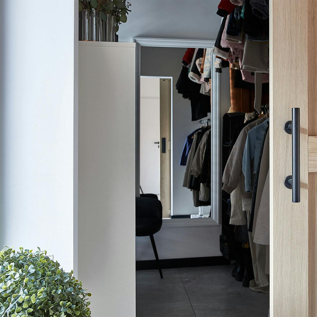 Detal dębowych drzwi przesuwnych, które uchylone odsłaniają wnętrze garderoby. Wewnątrz widać ubrania wiszące na wieszakach i lustro