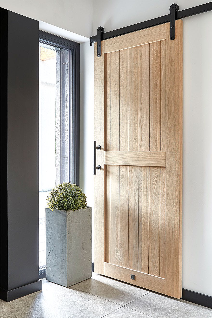 A view of the oak sliding doors in a modern vestibule. The light, natural wood door with a black sliding system and handle is the focal point of the design, next to which sits a large concrete planter.