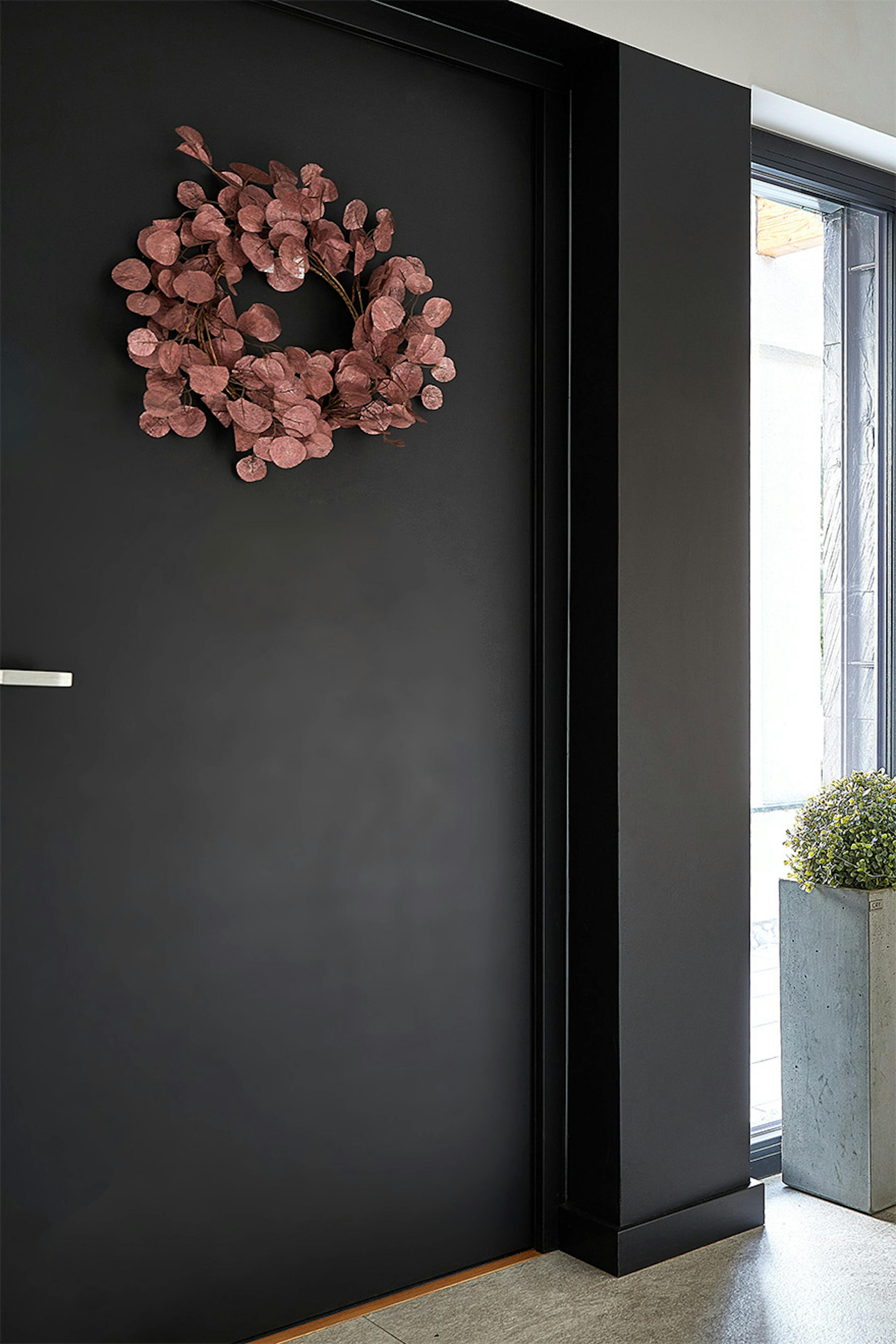 Part of the vestibule with a black door, on which hangs a wreath of dried pink leaves. Next to it is a large concrete planter with a green plant, creating a contrasting color.