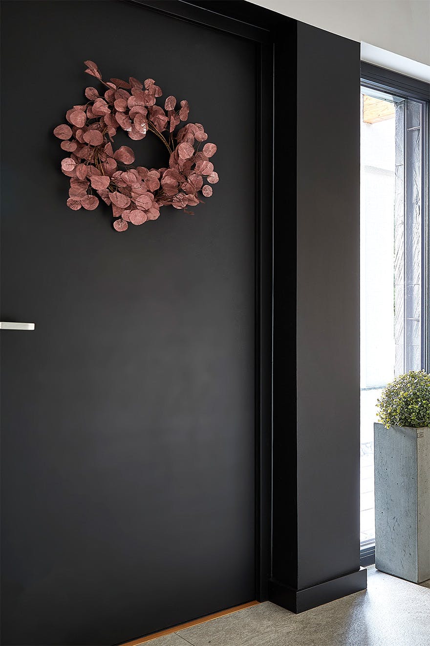 Part of the vestibule with a black door, on which hangs a wreath of dried pink leaves. Next to it is a large concrete planter with a green plant, creating a contrasting color.