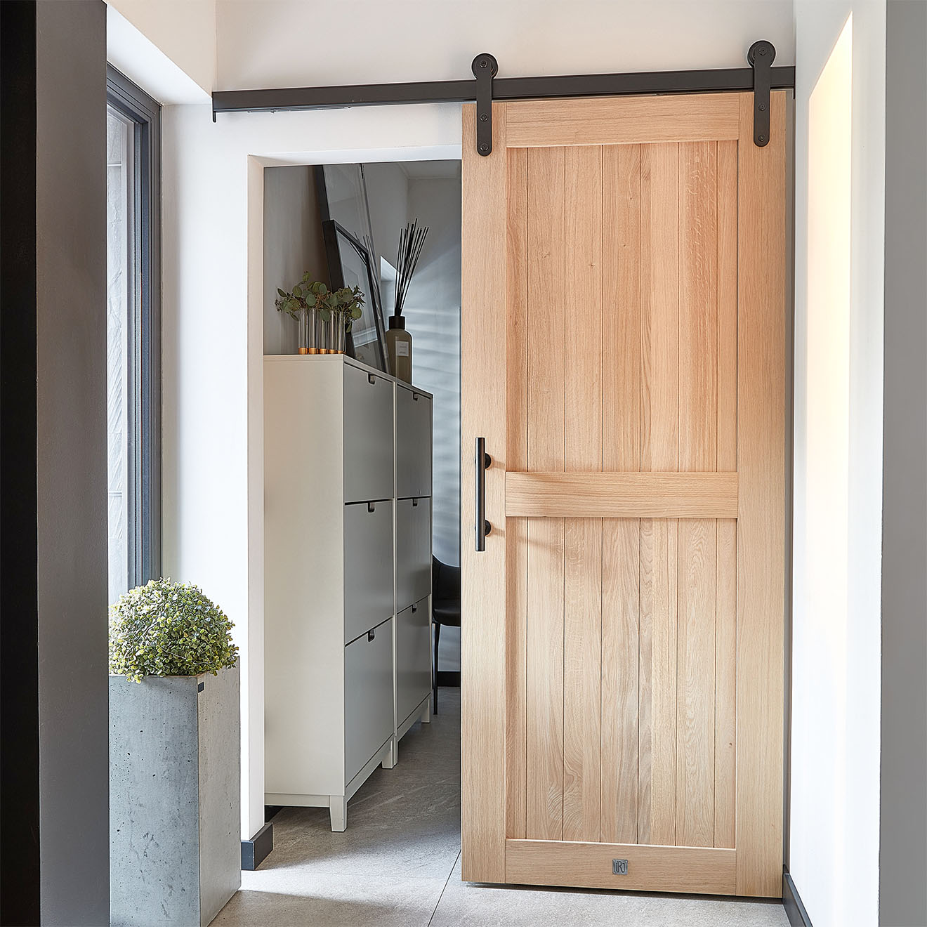 Ein eleganter Windfang mit Holzschiebetüren, die zu einem begehbaren Ankleide führen. Türen aus Natureiche mit schwarzen Beschlägen, ein großer Blumentopf und ein Schuhschrank schaffen einen funktionalen und stilvollen Raum.