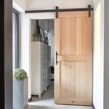 Ein eleganter Windfang mit Holzschiebetüren, die zu einem begehbaren Ankleide führen. Türen aus Natureiche mit schwarzen Beschlägen, ein großer Blumentopf und ein Schuhschrank schaffen einen funktionalen und stilvollen Raum.