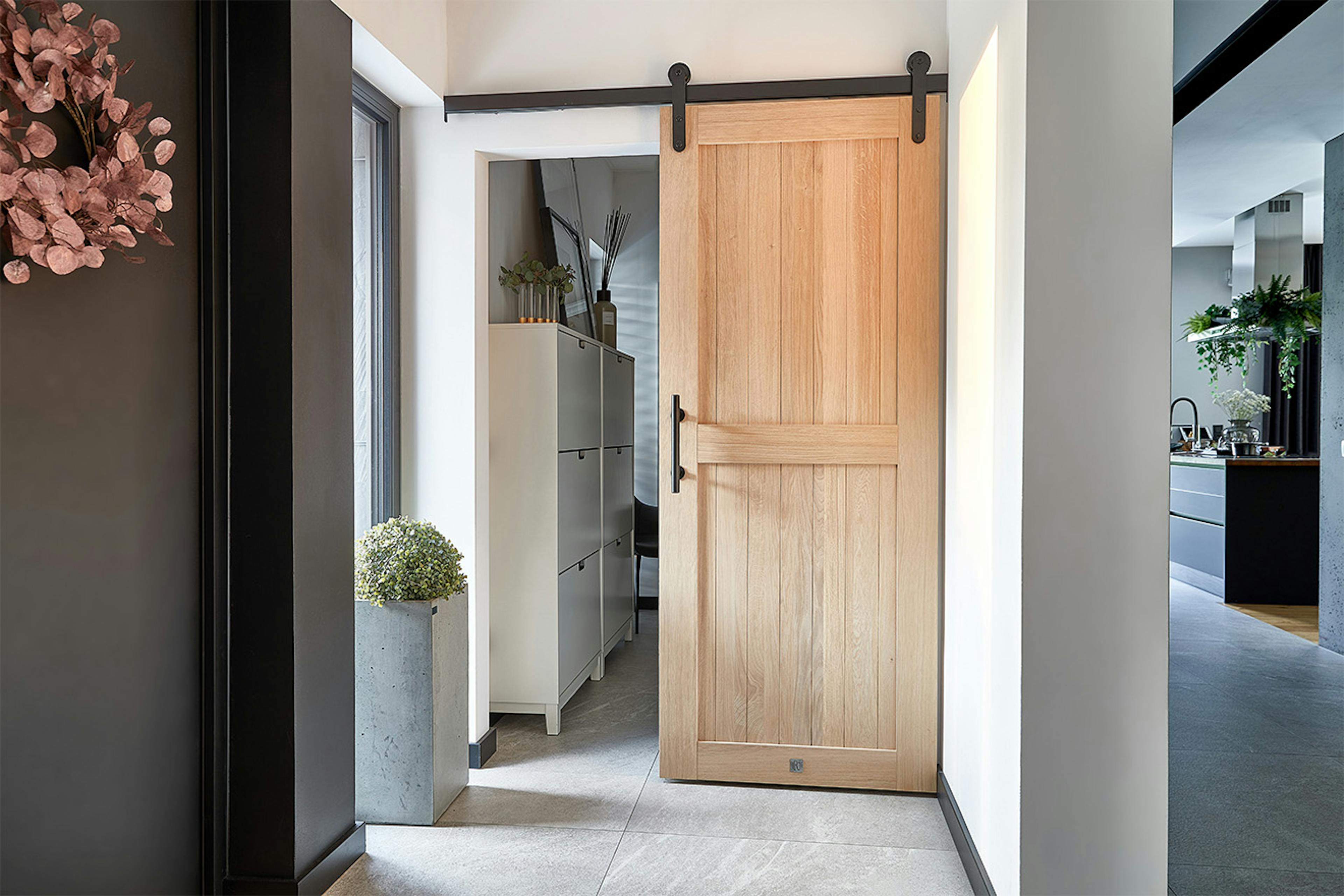 Portes coulissantes en chêne dans un vestibule moderne. Une porte de style grange en bois clair, suspendue à un rail noir, ouvre sur un dressing avec une armoire à chaussures blanche.