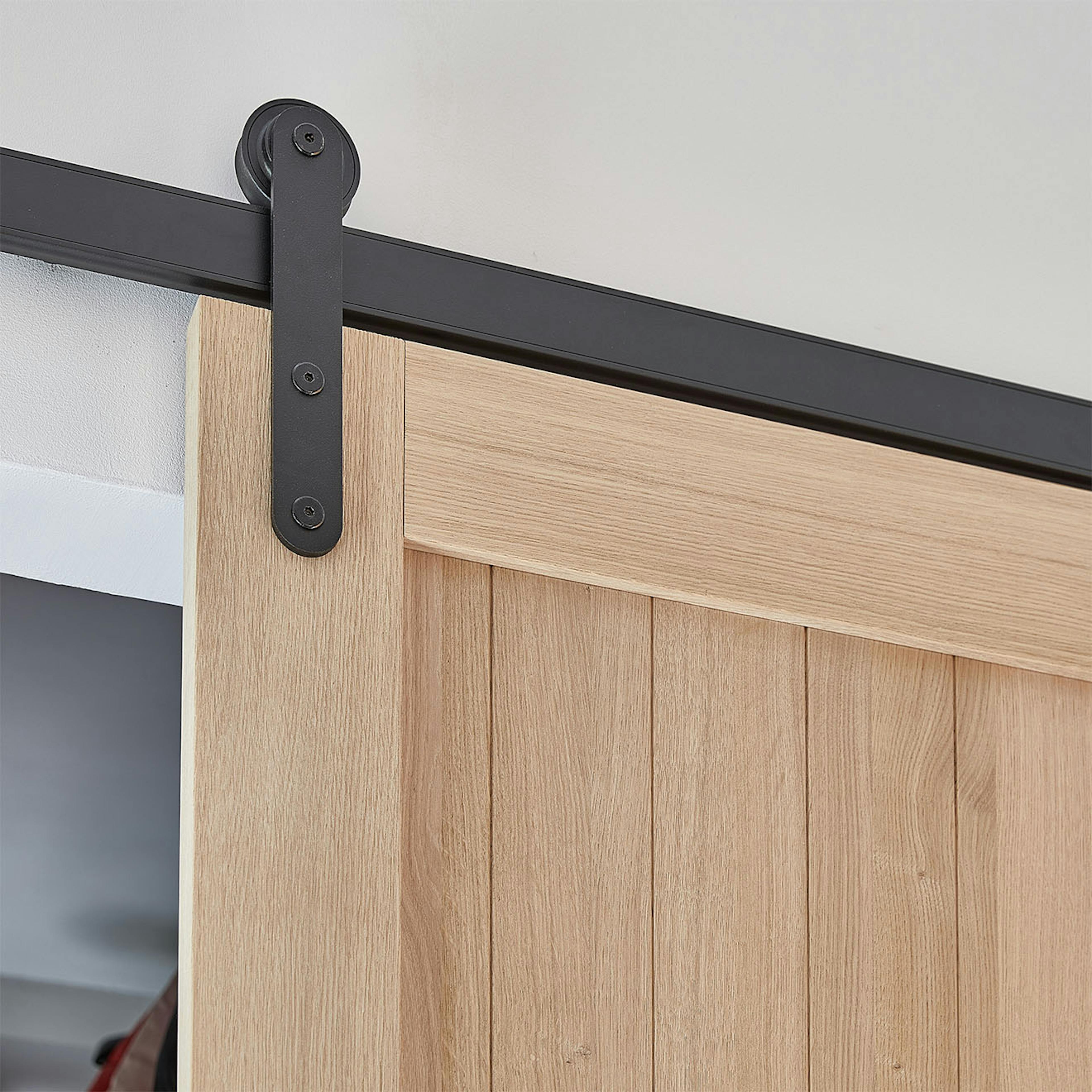 Gros plan sur le système coulissant noir (ferrures) et la partie supérieure de la porte en chêne. Le système noir contraste avec le bois clair et naturel de la porte, créant un mélange harmonieux de style industriel et naturel.