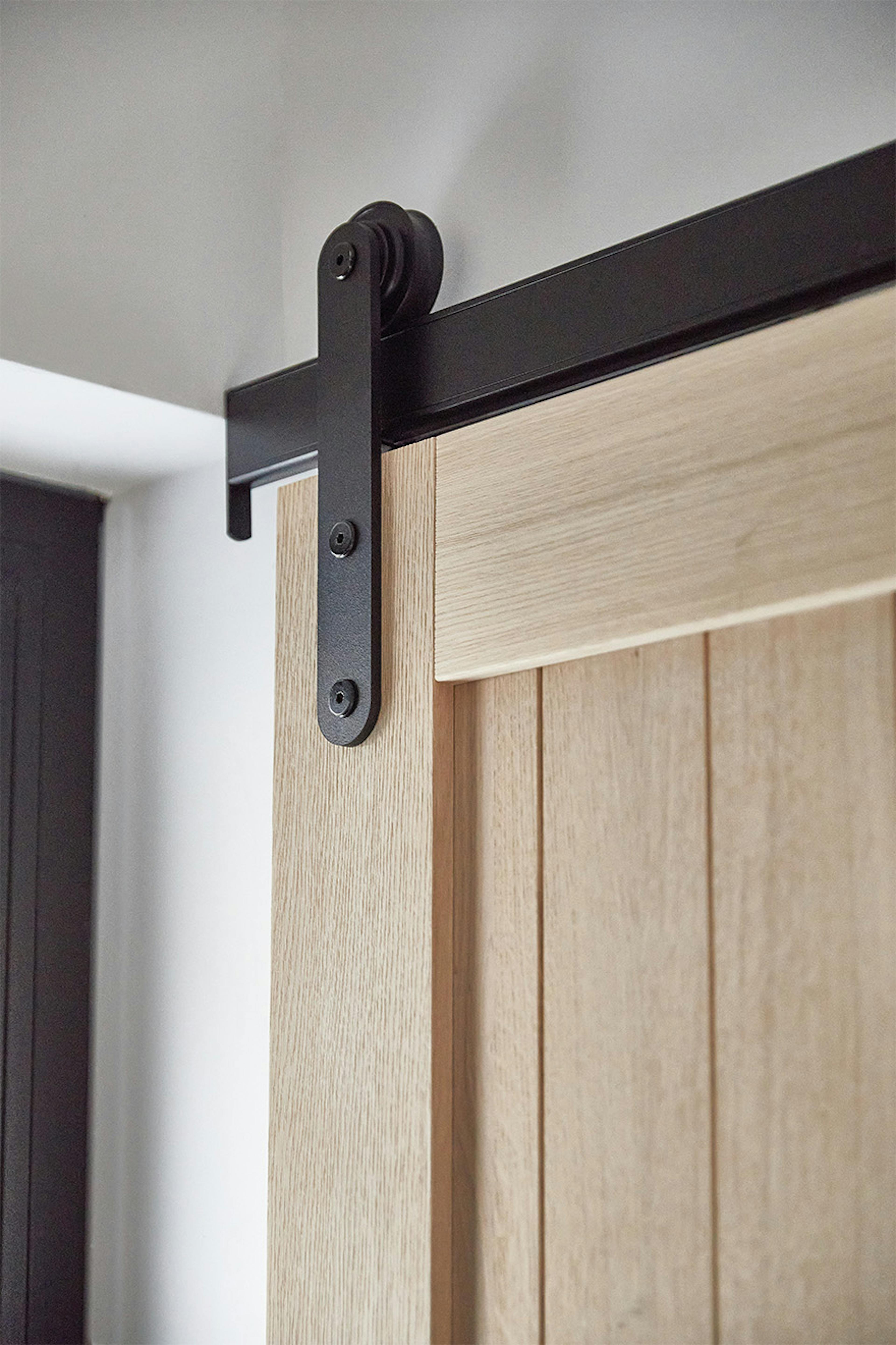 Gros plan sur le système coulissant noir (ferrures) et la partie supérieure de la porte en chêne. Le système noir contraste avec le bois clair et naturel de la porte, créant un mélange harmonieux de style industriel et naturel.
