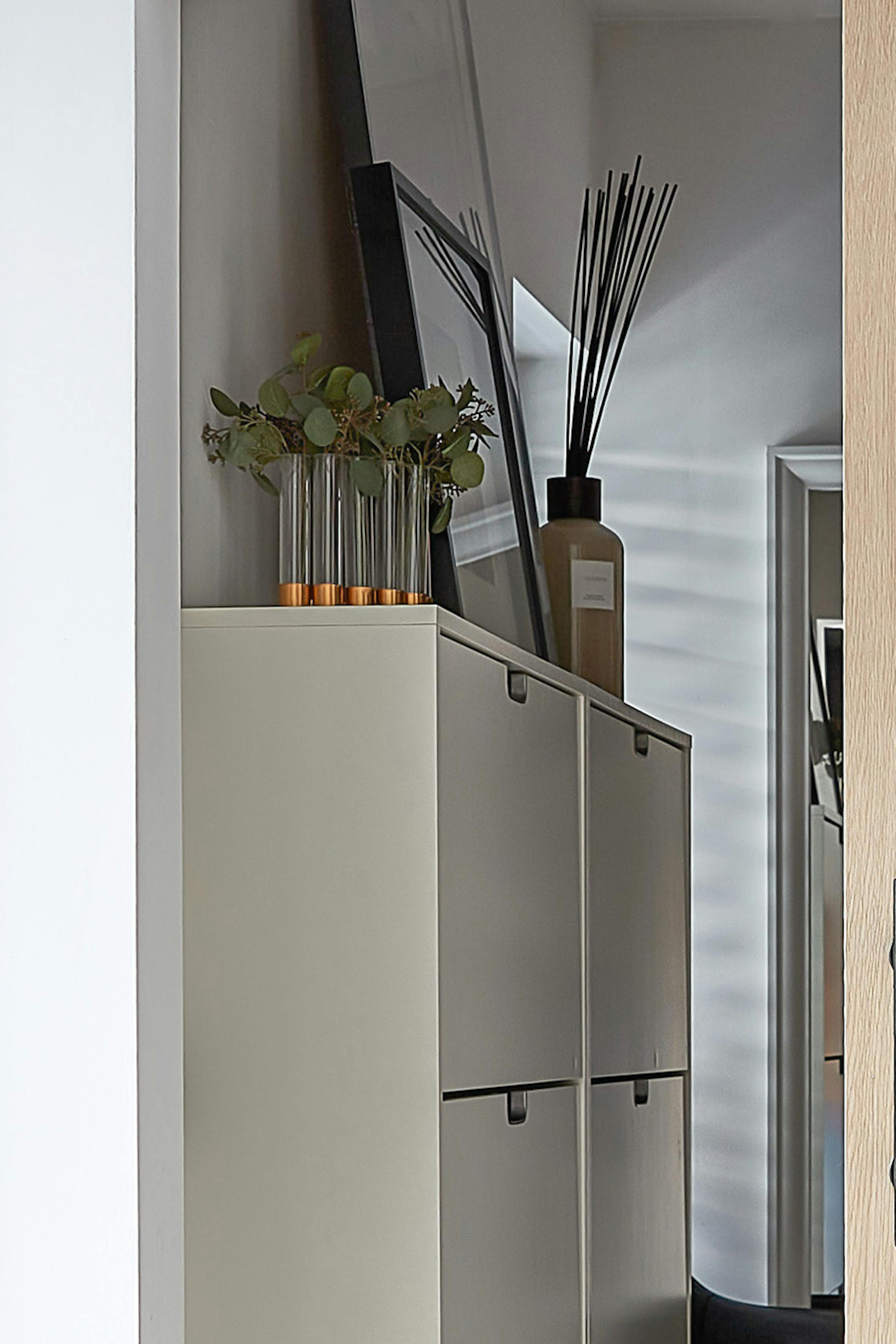 Détails de l'intérieur du dressing, visibles à travers la porte en chêne entrouverte. Des décorations élégantes trônent sur un meuble à chaussures blanc : un vase d'herbes aromatiques et un diffuseur de parfum, conférant à l'espace une atmosphère chaleureuse et élégante.