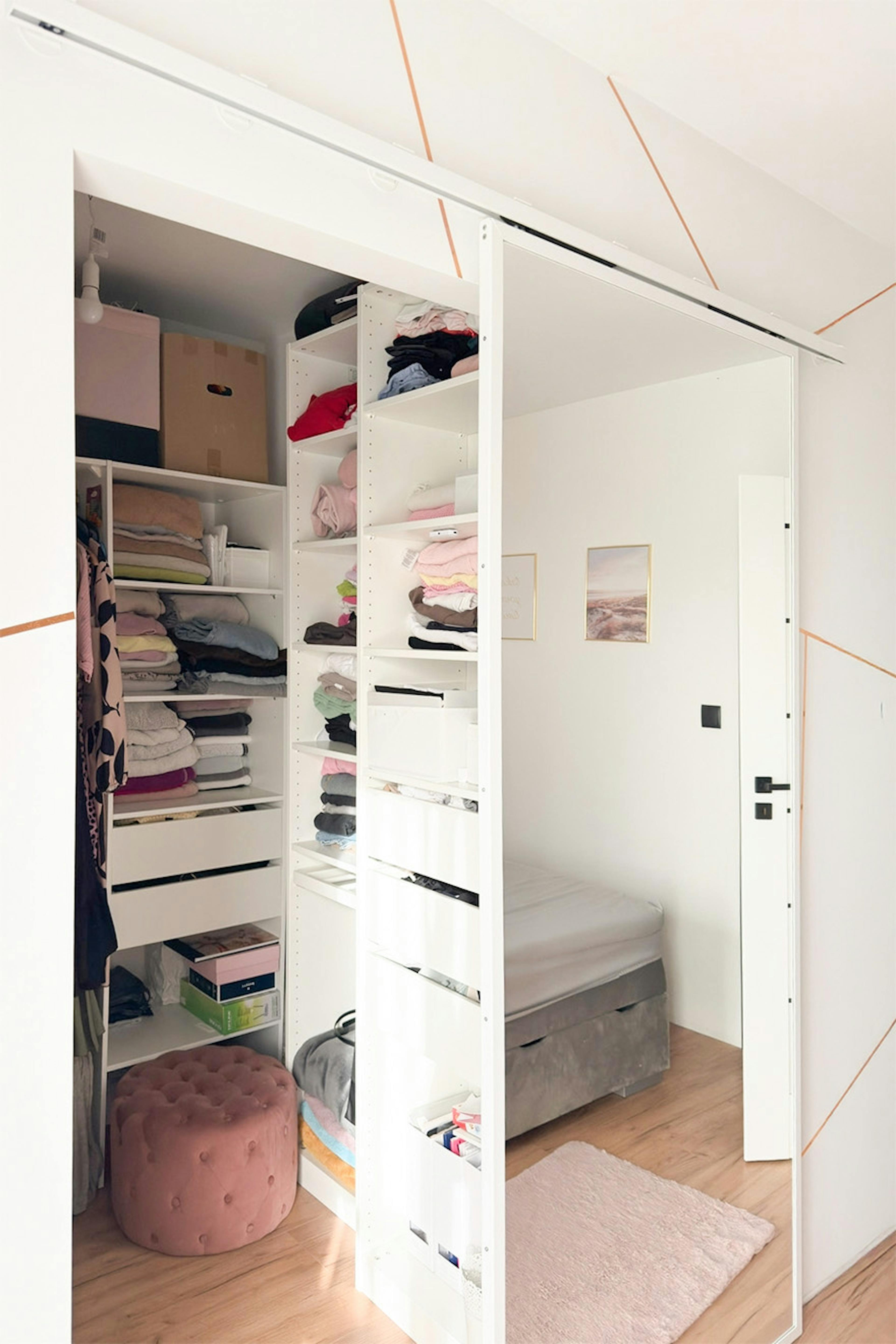 Penderie ouverte blanche avec étagères et tiroirs ; reflet du lit dans le miroir ; pouf en velours couleur poudre dans la penderie.