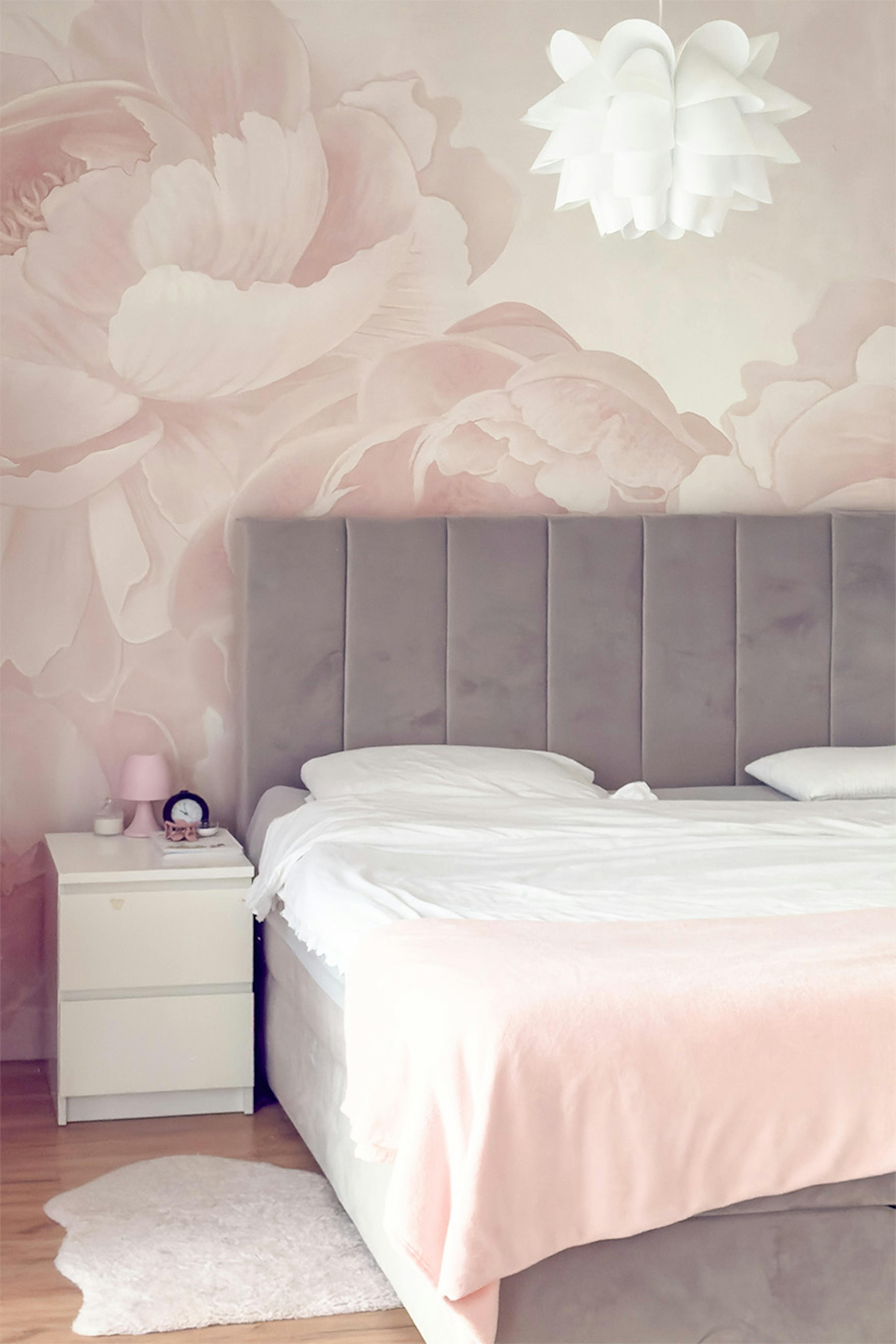 Lit avec tête de lit matelassée grise ; papier peint avec de grandes fleurs roses dans la chambre ; table de chevet blanche.