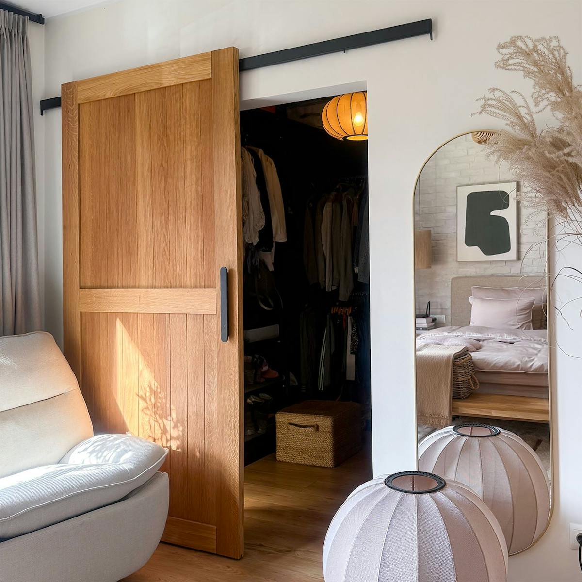 Offene Schiebetüren aus Eichenholz führen von einem hellen, beigen Schlafzimmer in einen praktischen begehbaren Kleiderschrank. Im Raum fallen ein bequemer Sessel und eine stimmungsvolle Beleuchtung ins Auge.